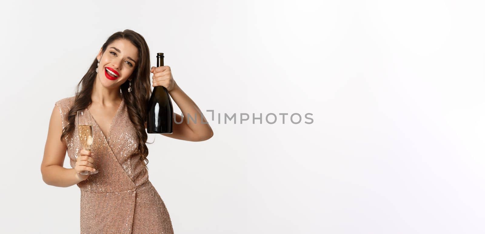 Winter holidays celebration concept. Happy woman on Christmas party saying cheers, raising bottle of champagne and smiling, standing in elegant dress, white background.