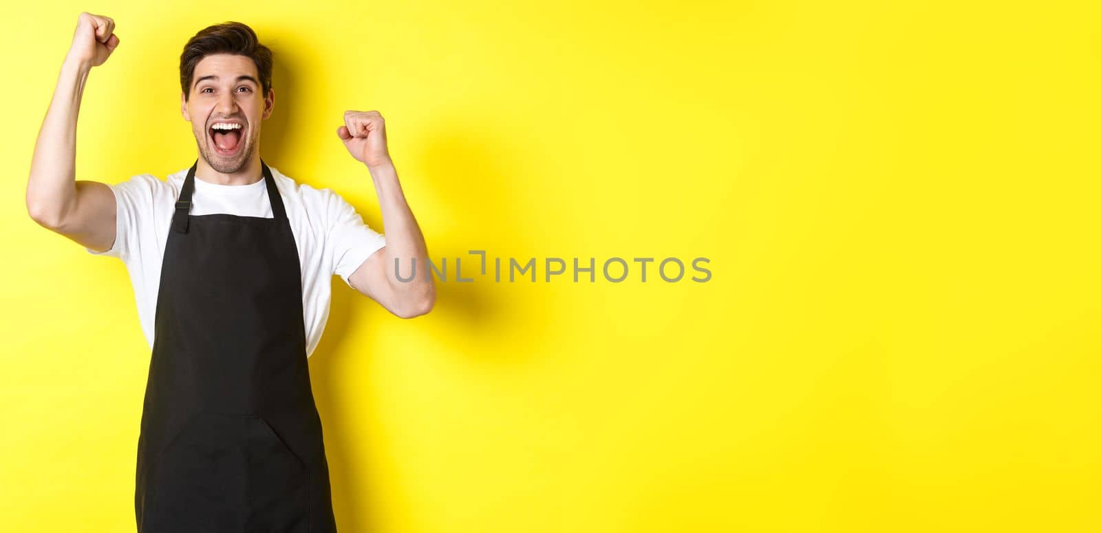 Happy barista celebrating victory, raising hands up and shouting for joy, wearing black apron, shop uniform, standing against yellow background.