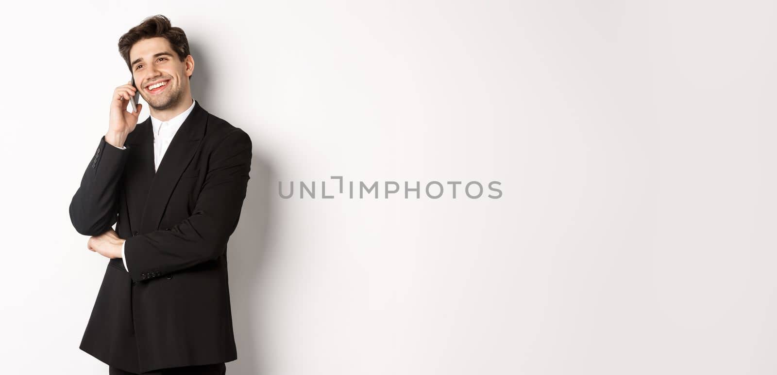 Image of handsome and successful businessman talking on phone, smiling pleased, standing in suit against white background.