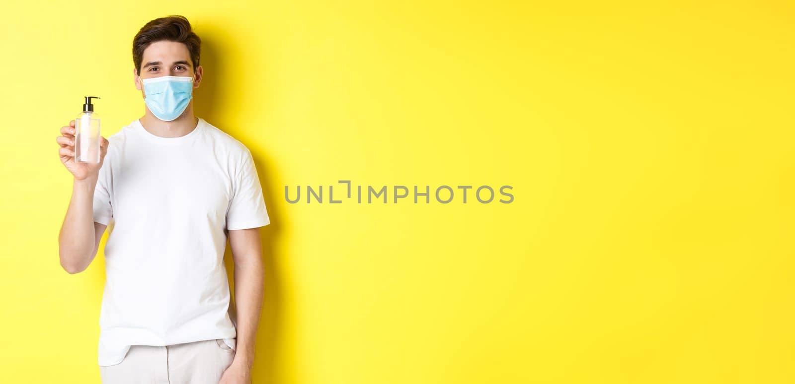 Concept of covid-19, quarantine and lifestyle. Young man in medical mask showing hand sanitizer, hands disinfection product, standing over yellow background.