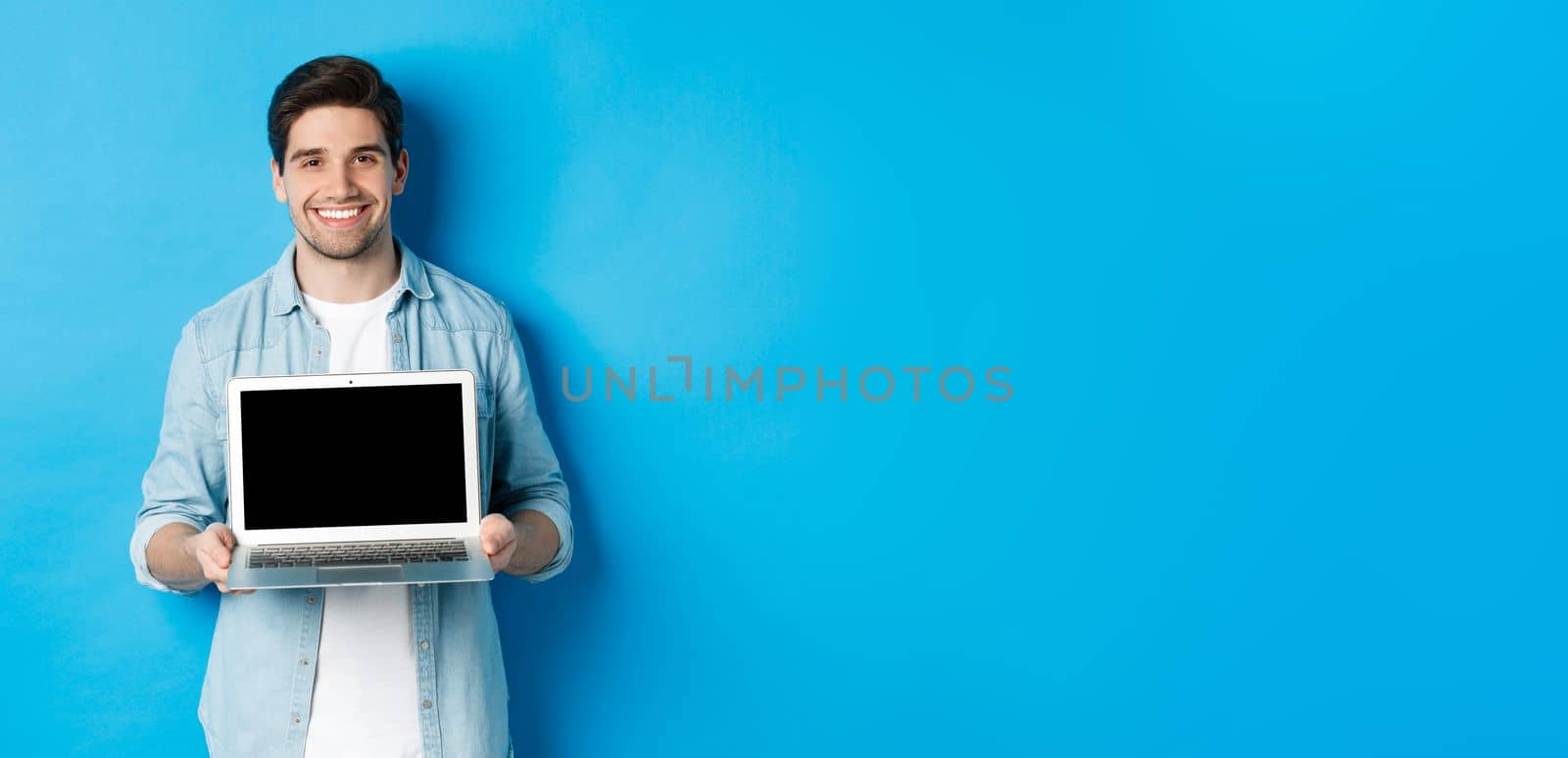 Handsome young man introduce product on laptop screen, showing computer and smiling, standing over blue background by Benzoix