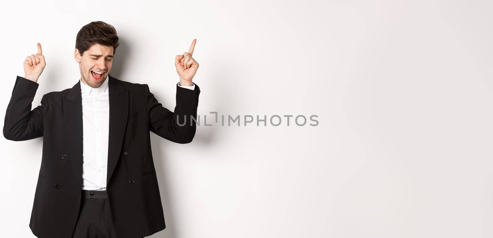 Image of successful handsome businessman dancing, enjoying party, pointing fingers up and having fun, standing over white background, wearing black suit.