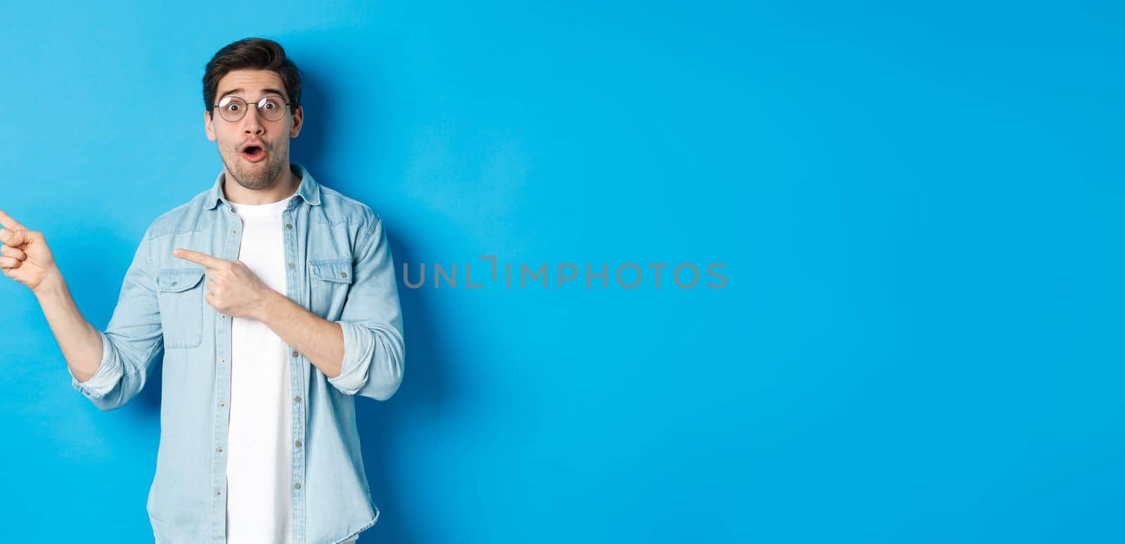 Surprised man in glasses pointing fingers left at copy space, asking question about product, looking intrigued, standing over blue background.