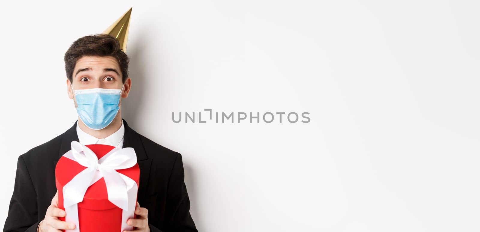 Concept of party during covid-19. Close-up of happy guy in trendy suit and medical mask, celebrating christmas, holding present and smiling, standing over white background by Benzoix