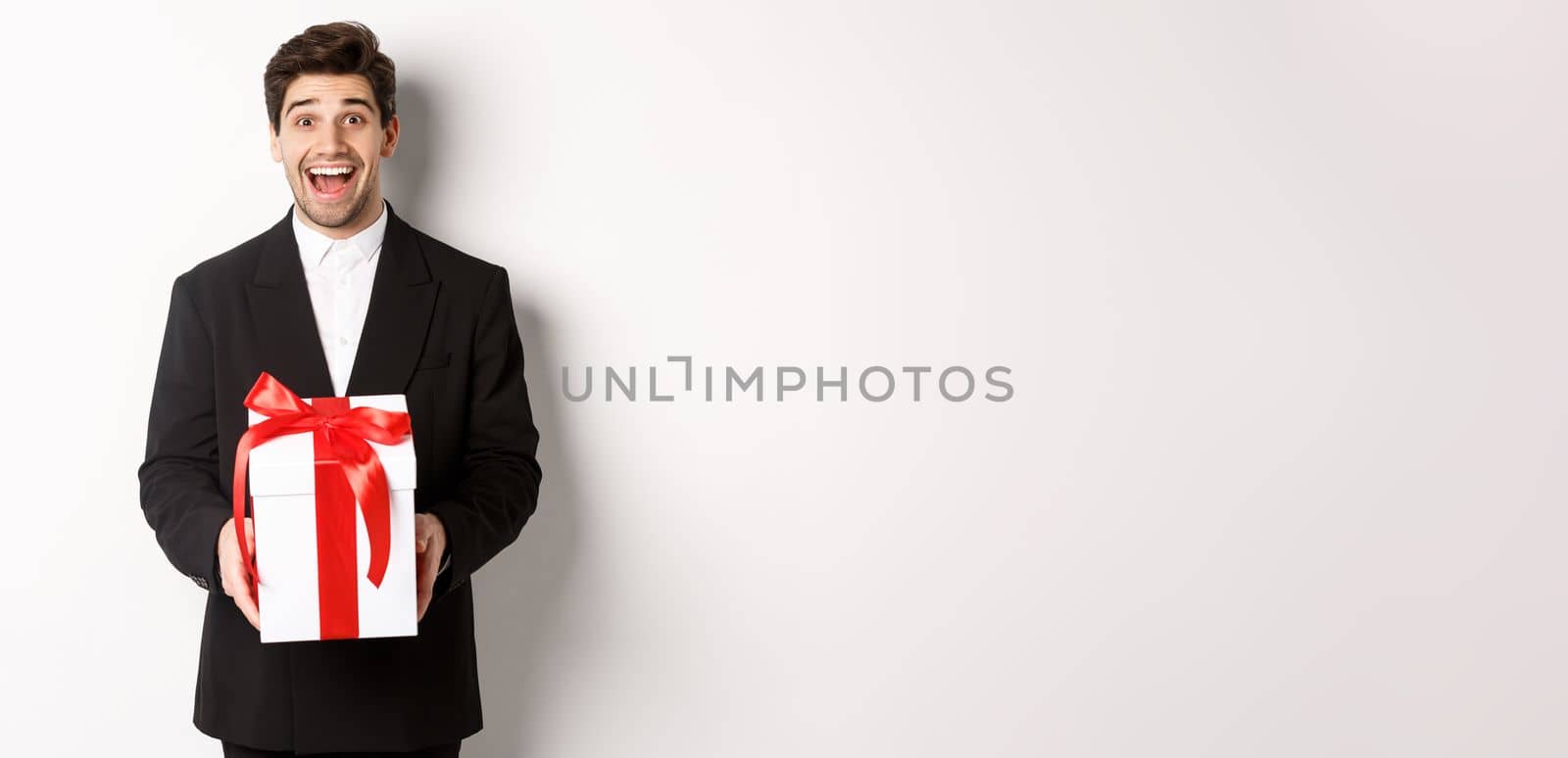 Concept of christmas holidays, celebration and lifestyle. Image of handsome guy in black suit looking excited, have a gift, standing against white background.