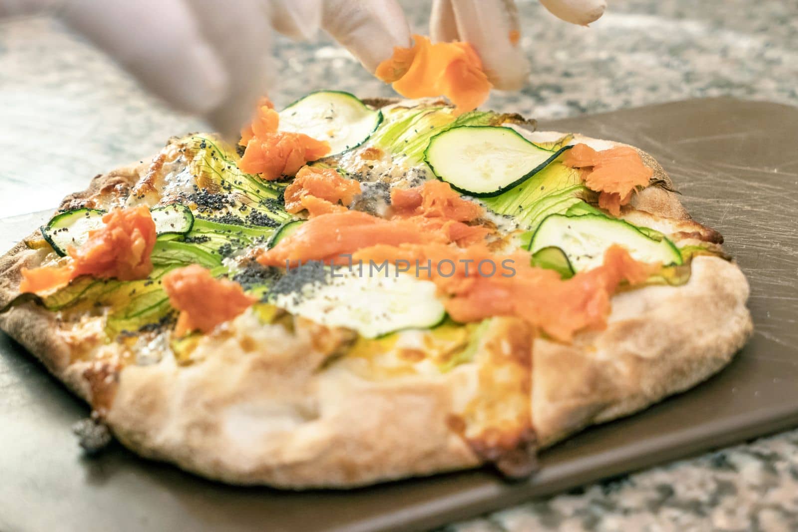 Delicious gourmet pizza with smoked salmon and courgette flowers. From above a hand with latex gloves decorates the pizza.