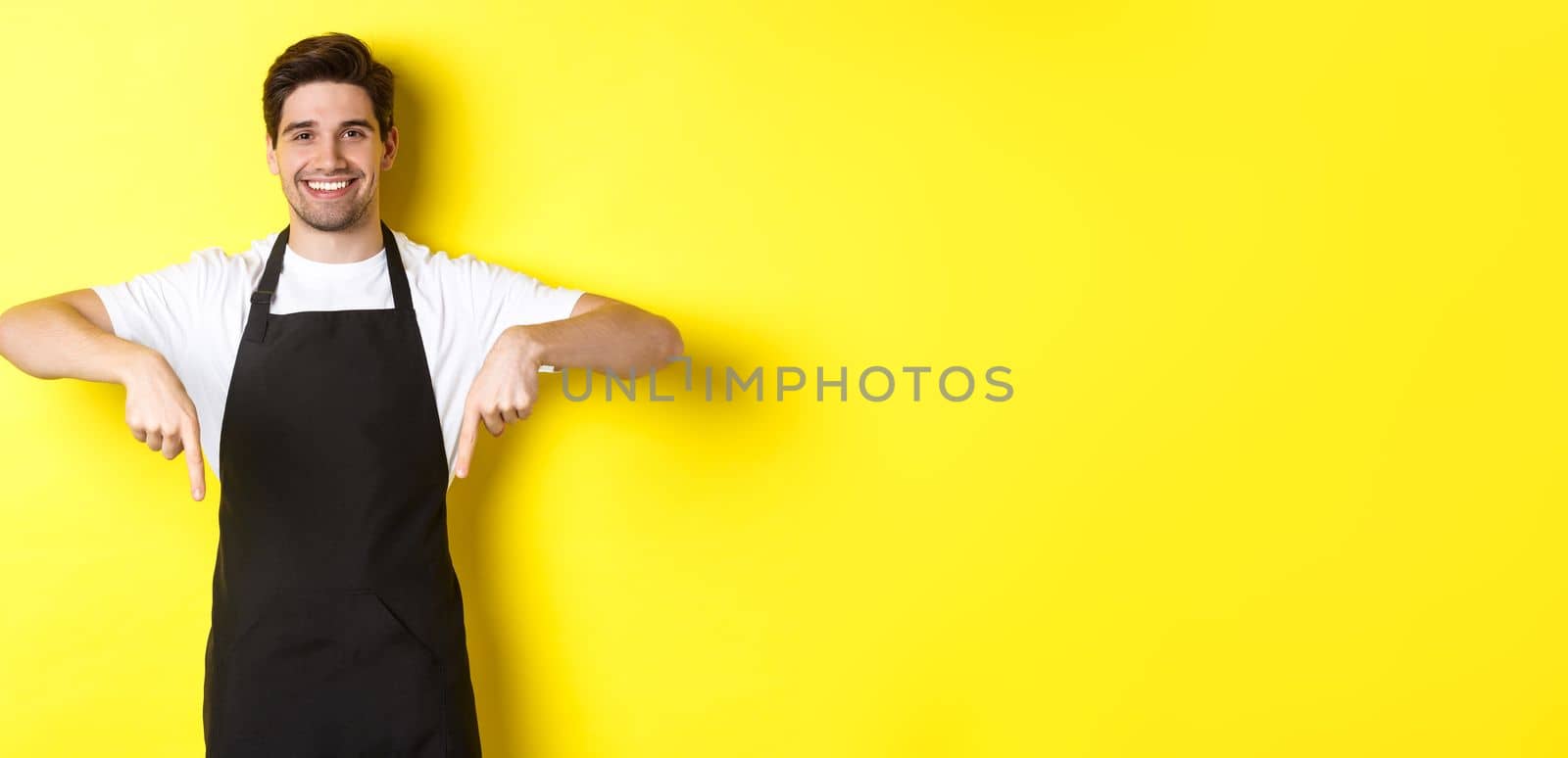 Friendly barista in black apron pointing fingers down, showing your logo banner, standing over yellow background.