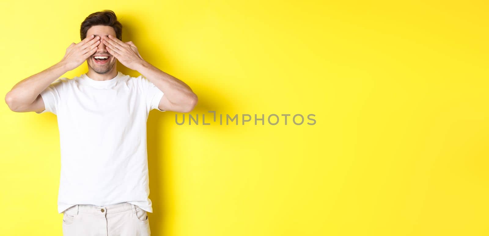 Happy man close eyes and waiting for surprise, smiling amused, standing over yellow background.