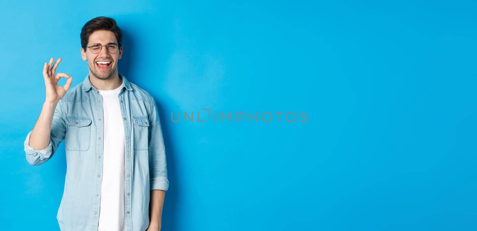 Confident smiling man in glasses showing ok sign, winking to guarantee or recommend something, blue background by Benzoix