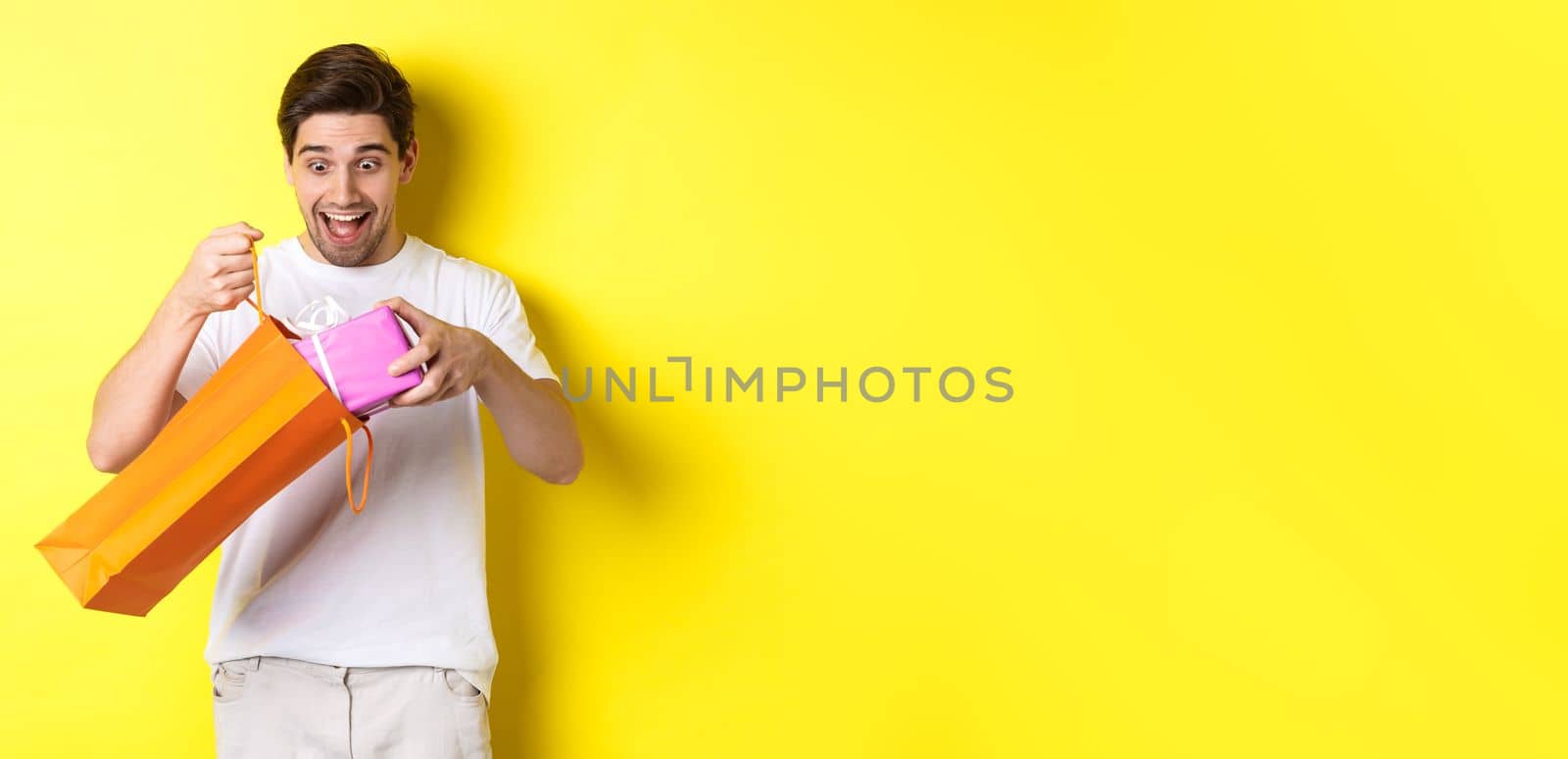 Concept of holidays and celebration. Young man looking surprised as take out gift from shopping bag, standing over yellow background.