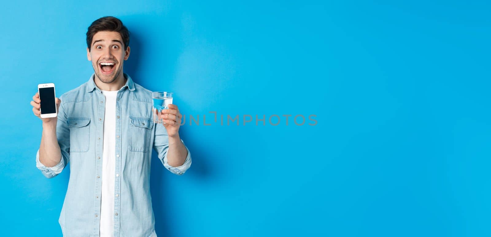 Happy man showing glass of water and mobile screen, recommending smartphone health app, standing over blue background.