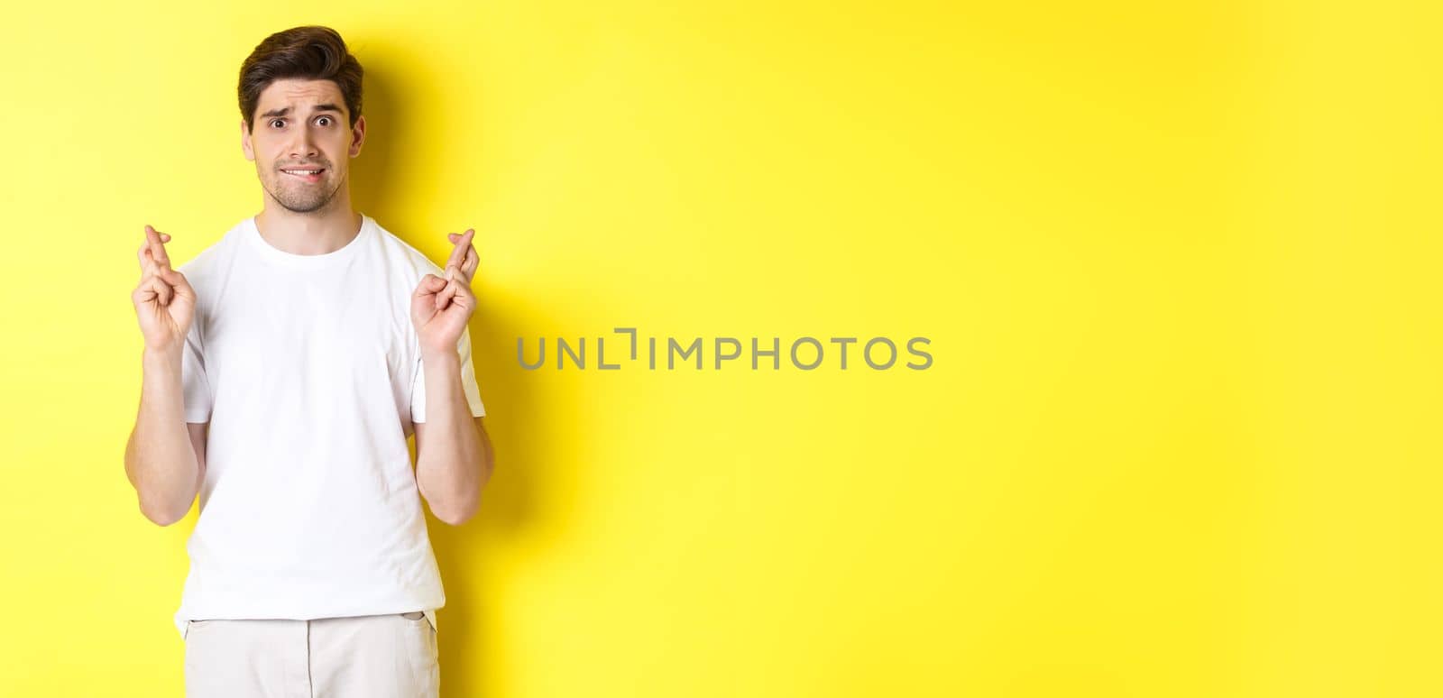 Nervous guy crossing fingers for good luck, hoping for something, standing over yellow background.