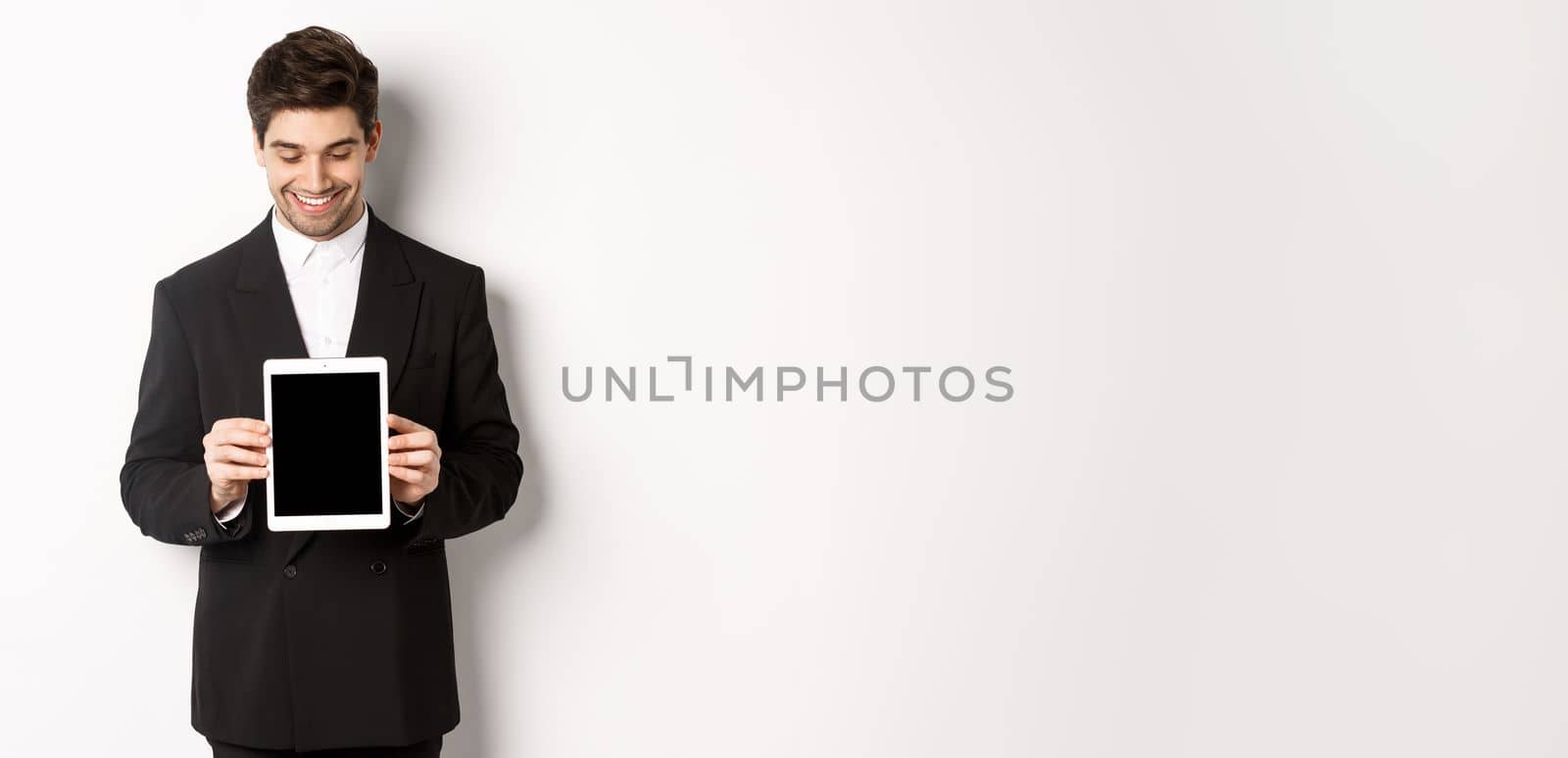 Image of good-looking male entrepreneur in black suit, looking down at digital tablet screen and showing advertisement, standing against white background.