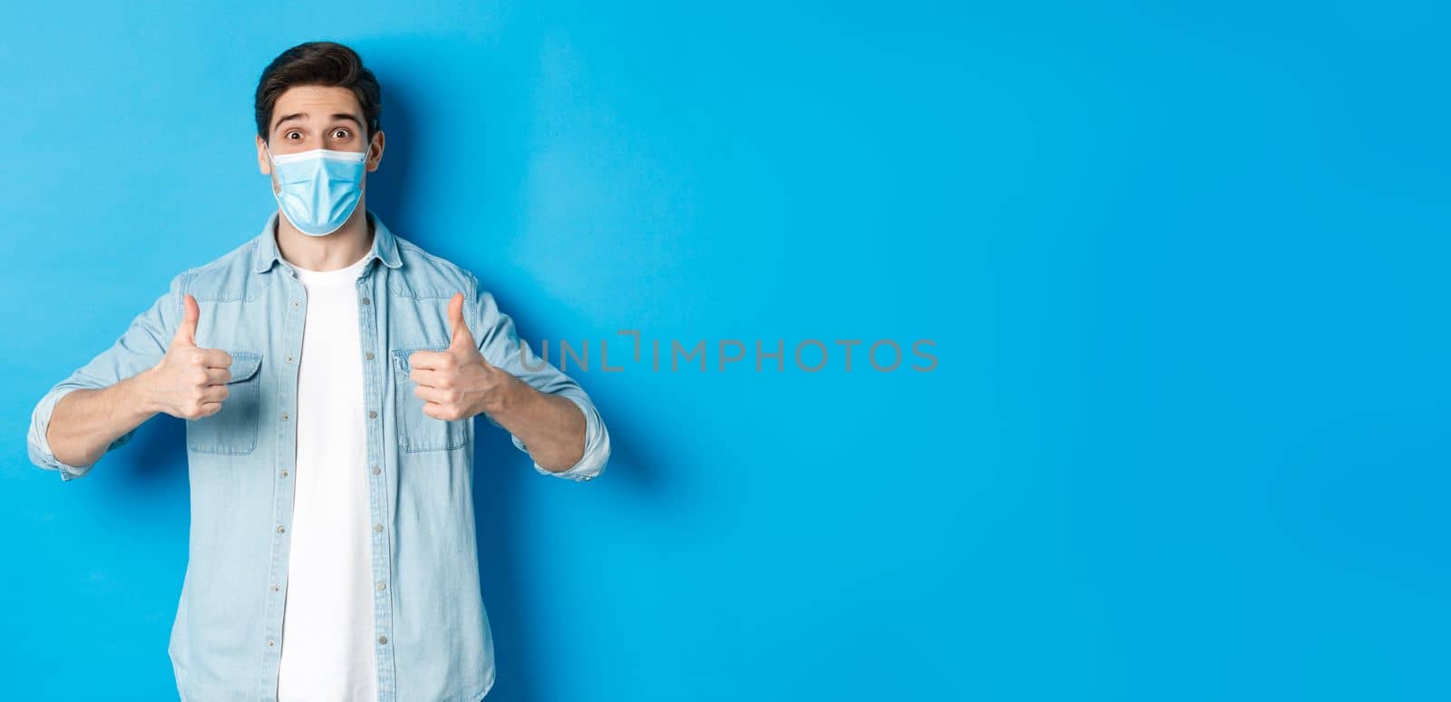 Concept of covid-19, pandemic and social distancing. Amazed guy in medical mask showing thumbs-up, recommending promo offer, standing against blue background.