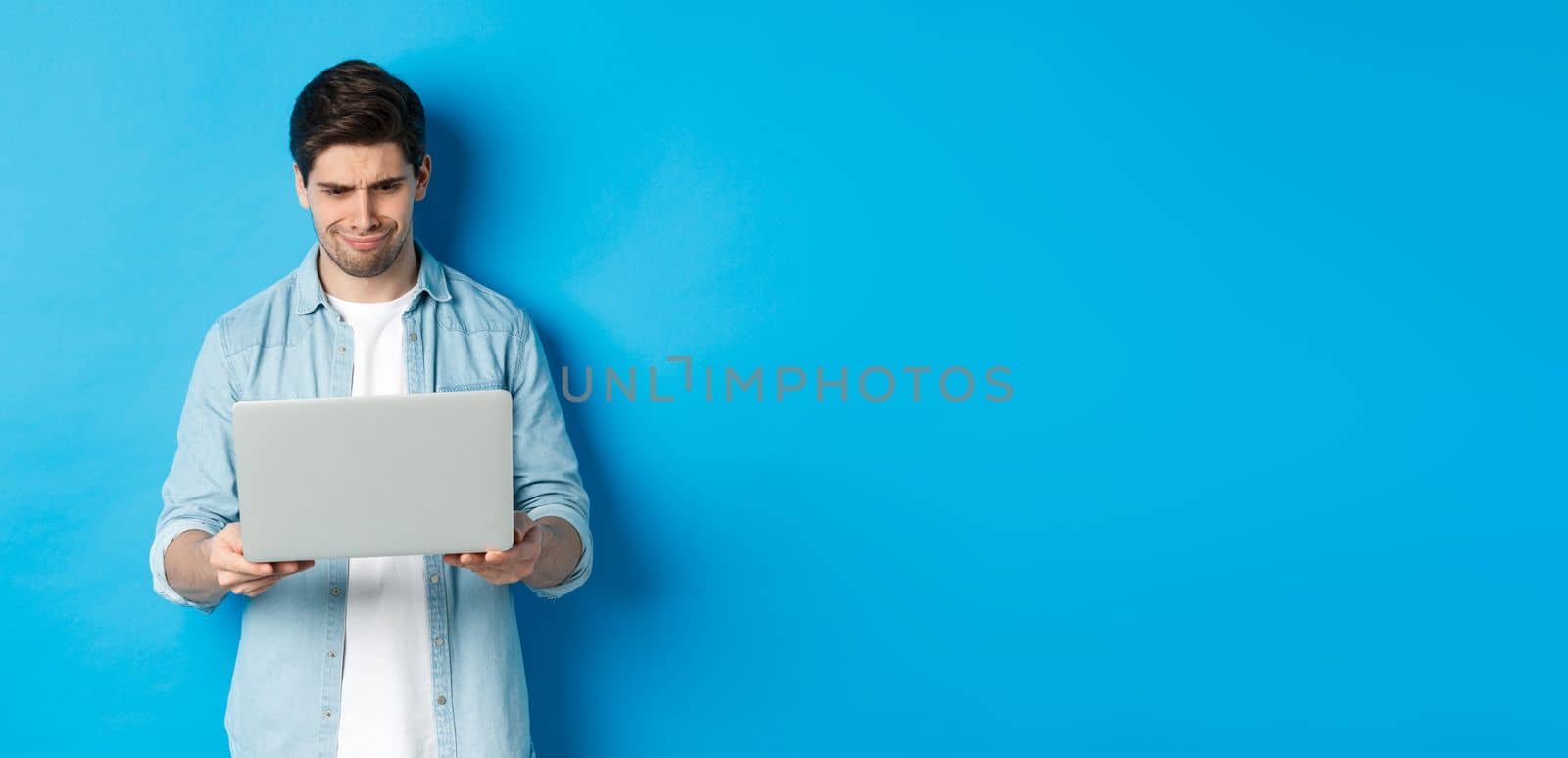 Disappointed handsome man looking at laptop screen and grimacing, judging something bad in internet, standing over blue background by Benzoix