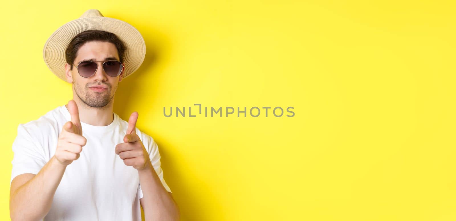 Concept of tourism and vacation. Close-up of cool guy in summer hat and sunglasses pointing finger guns at camera, standing over yellow background.