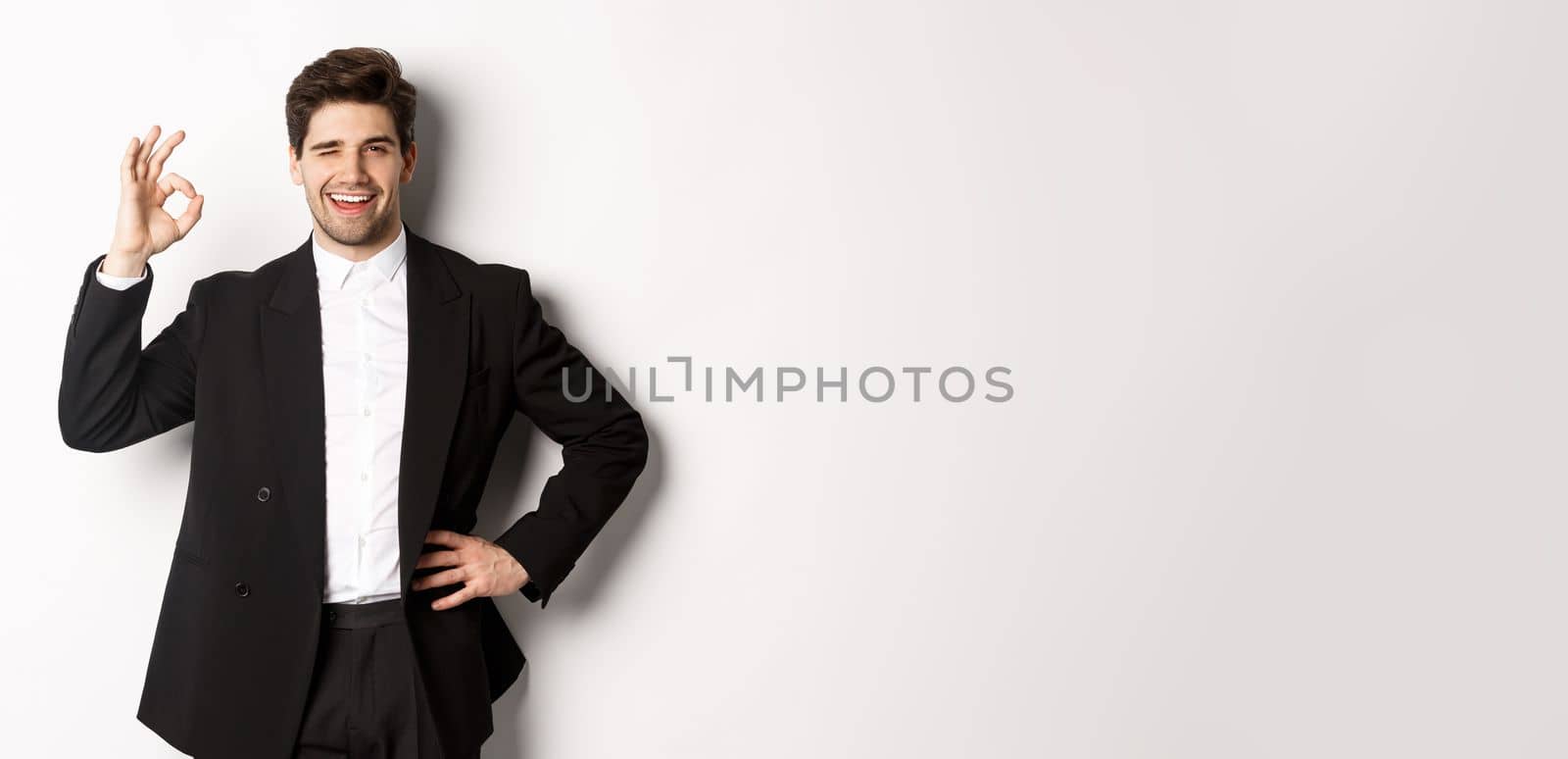 Concept of new year party, celebration and lifestyle. Portrait of confident, successful businessman in suit, showing okay sign and winking, approve something good, white background.
