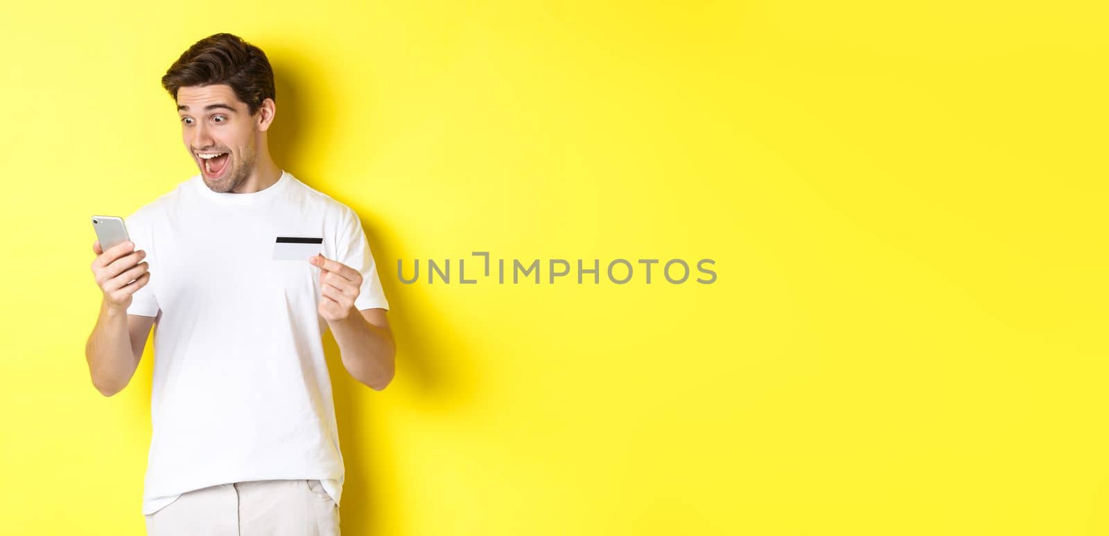 Man looking surprised at smartphone, shopping online, holding credit card, standing over yellow background by Benzoix