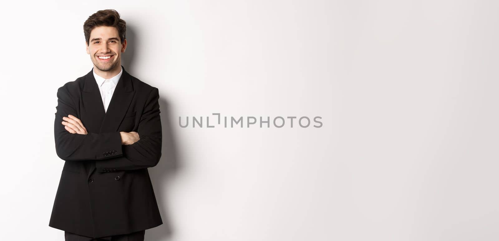 Handsome and confident bearded man, standing in formal suit with hands crossed on chest, smiling satisfied, attend party or work event, standing over white background.