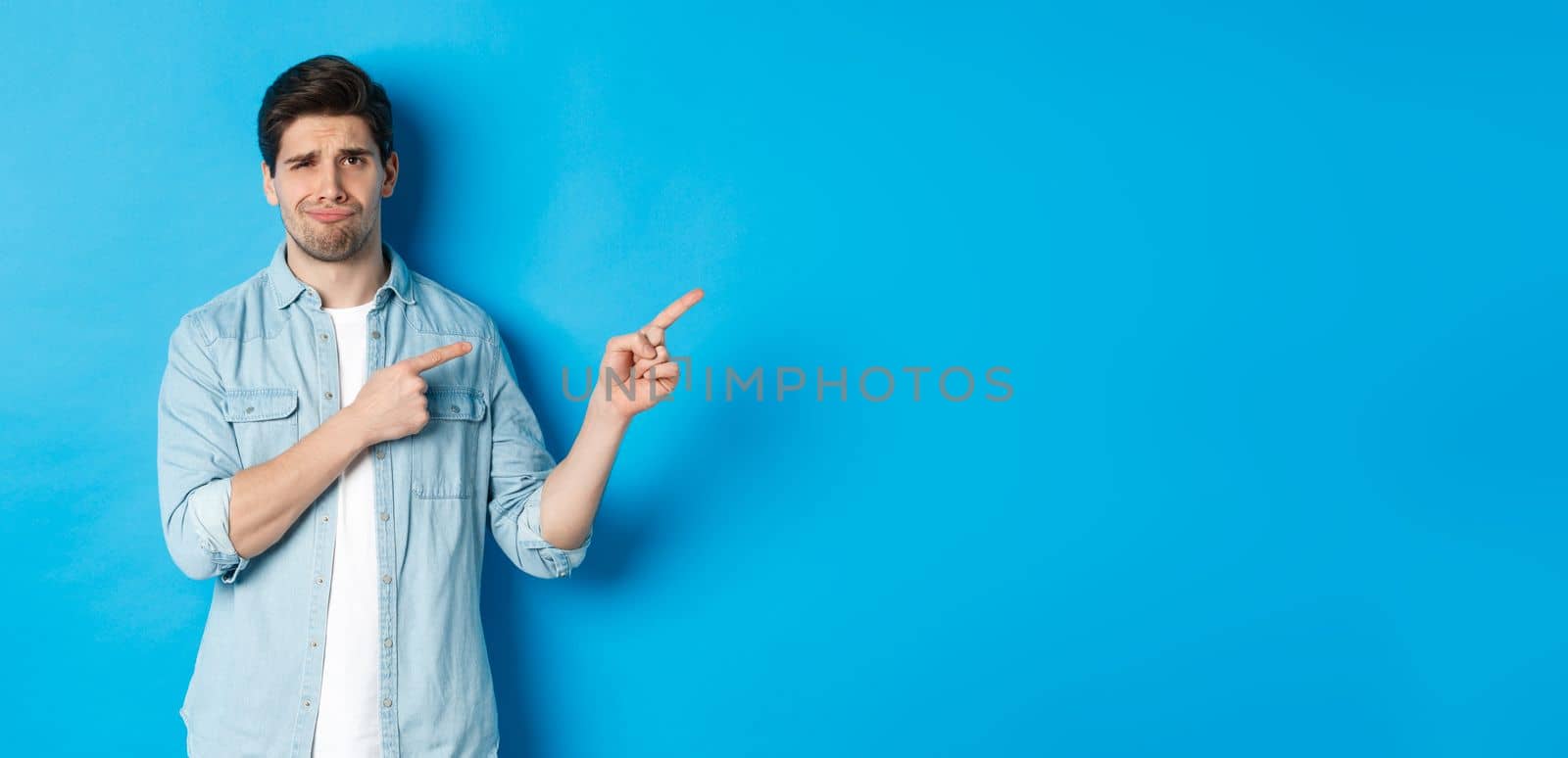 Portrait of skeptical adult guy pointing fingers right and smirking, exress disappointment and doubt, standing against blue background.