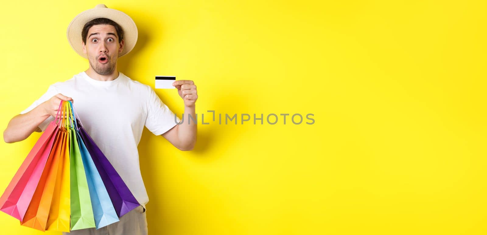 Impressed man showing shopping bags with products and credit card, standing over yellow background.