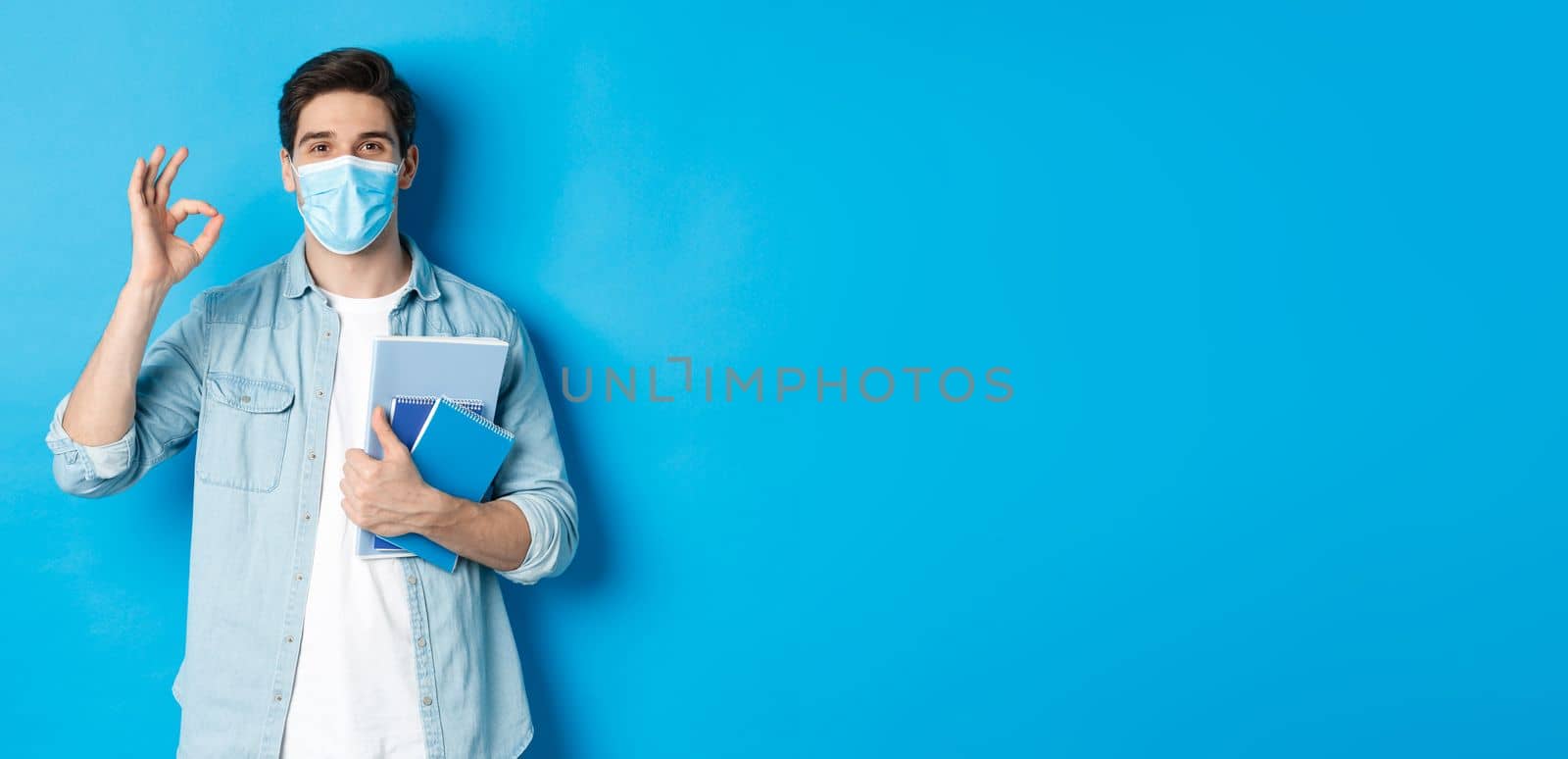 Education, covid-19 and social distancing. Guy student in medical mask looking happy, holding notebooks, showing ok sign, standing over blue background.