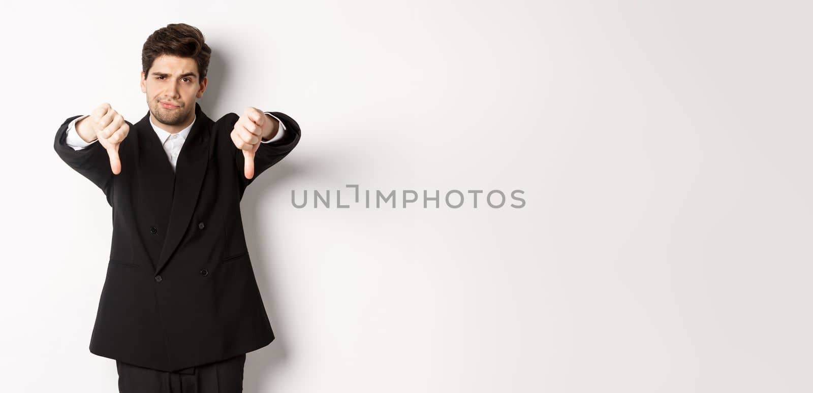 Portrait of skeptical and disappointed man in black suit, frowning upset, showing thumbs-down, dislike something bad, standing over white background by Benzoix