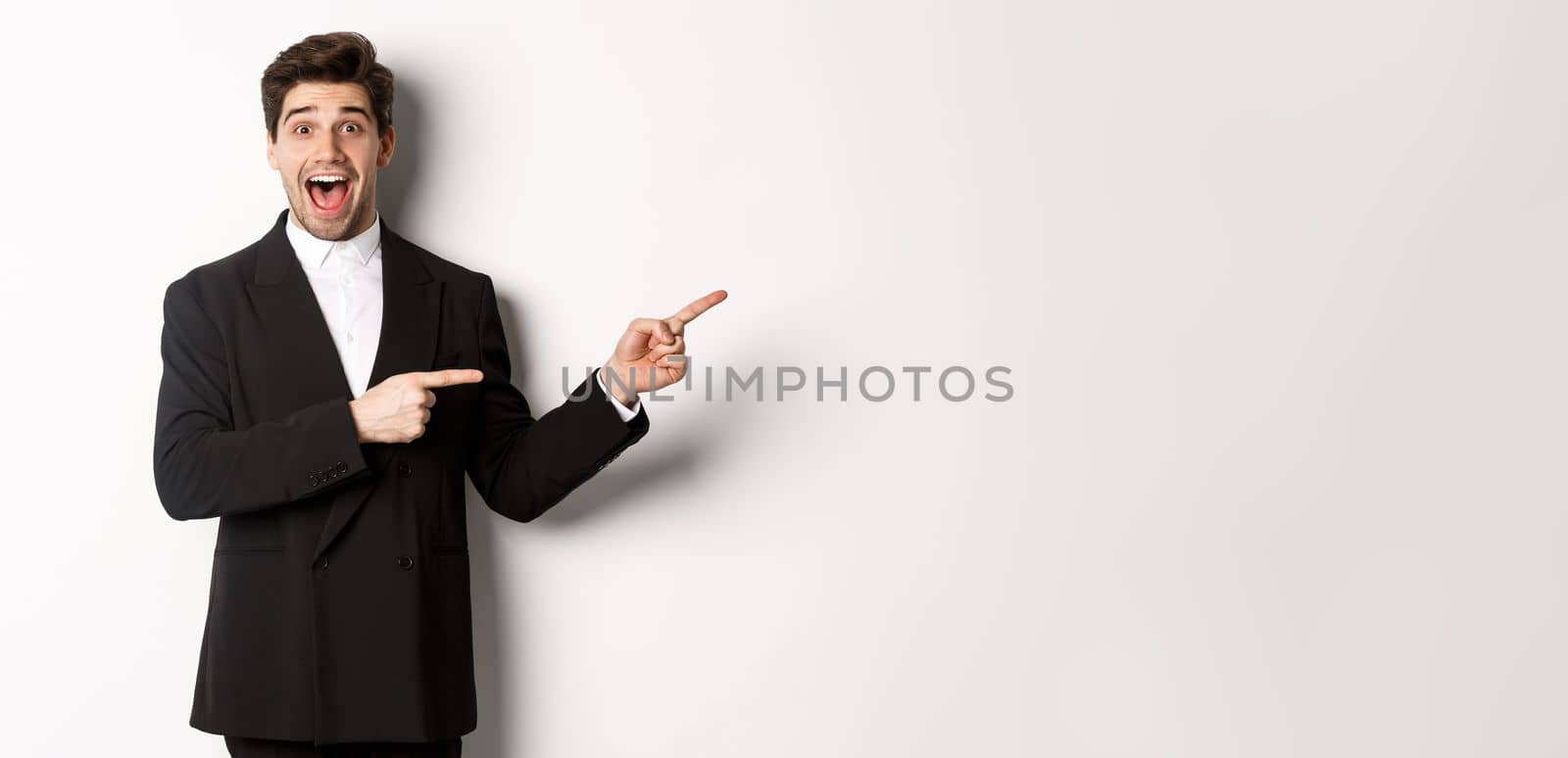 Image of attractive smiling guy dressed for new year party, pointing fingers right and showing advertisement, looking impressed standing over white background.