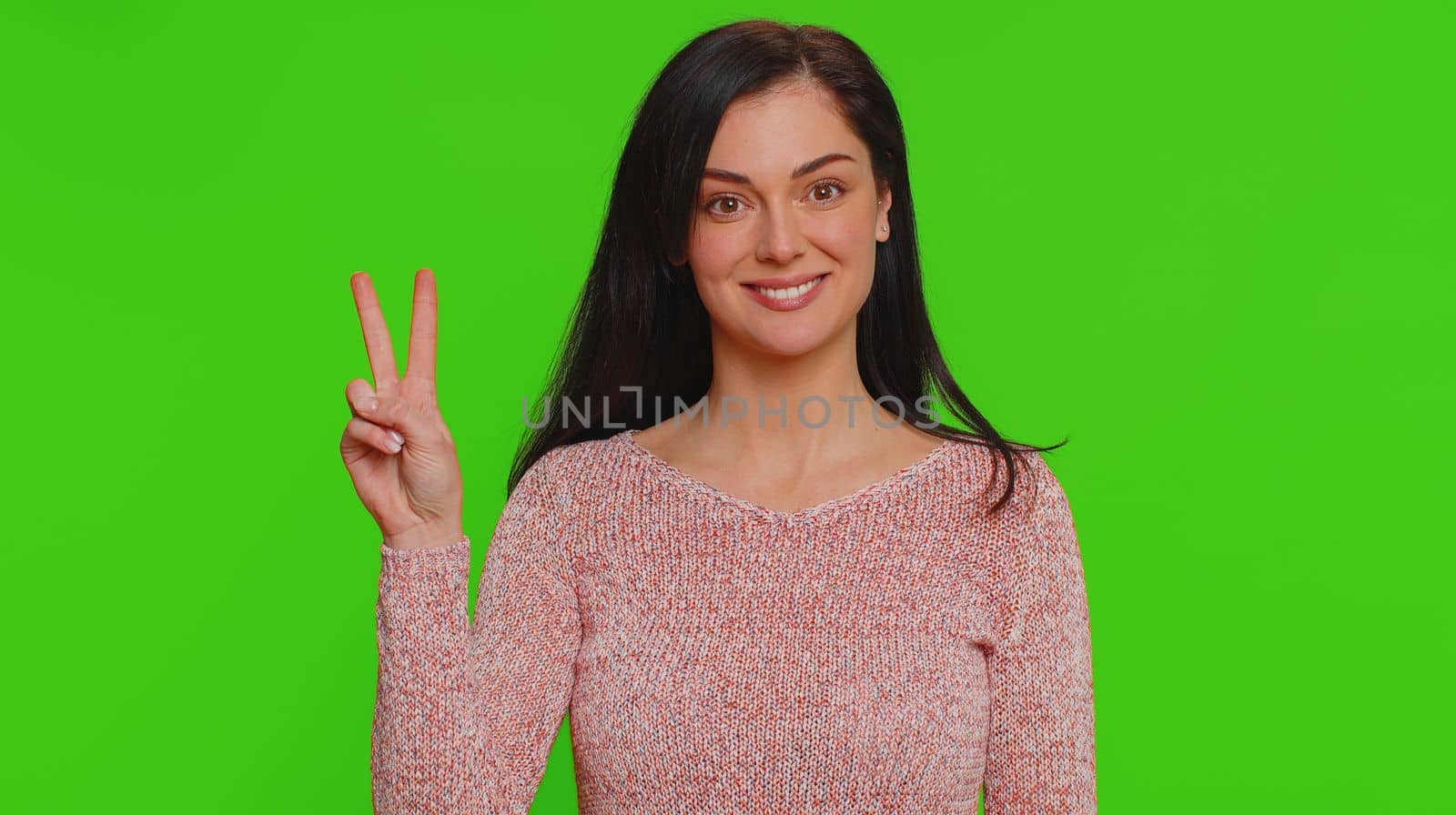 Hipster pretty woman in sweater showing victory v sign, hoping for success and win, doing peace gesture, smiling with kind optimistic expression. Young adult girl isolated on chroma key background