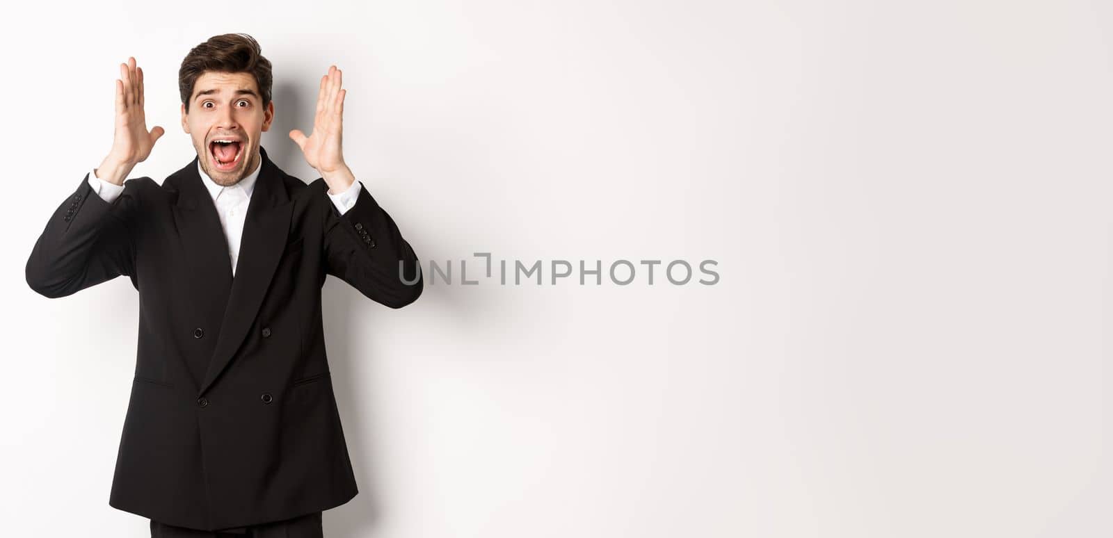 Frustrated and concerned man in black suit, screaming in panic and looking at something shocking, standing over white background.