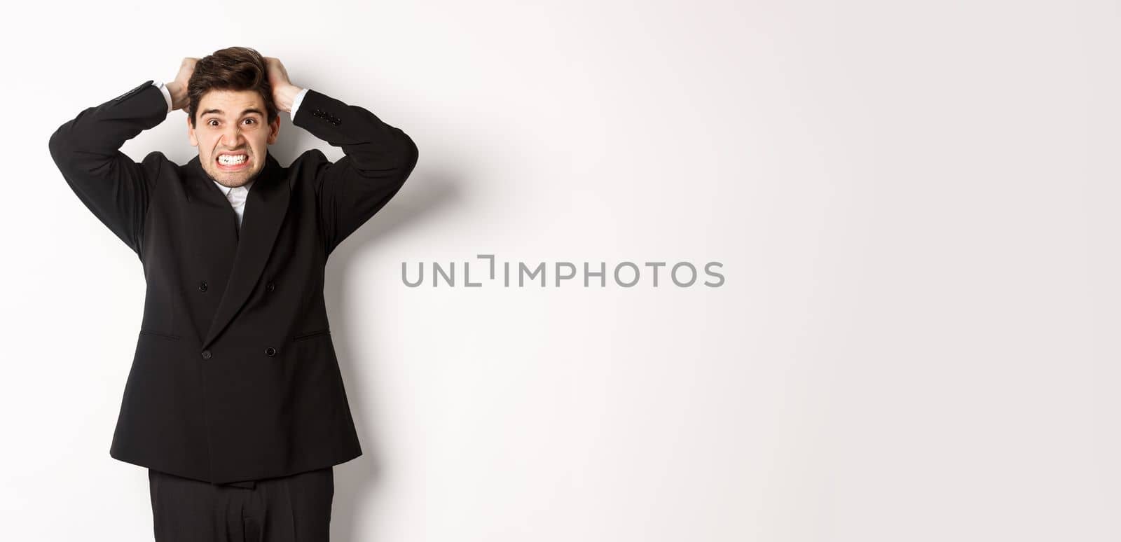 Image of frustrated and angry businessman in black suit, ripping hair on head and grimacing mad, standing tensed against white background by Benzoix