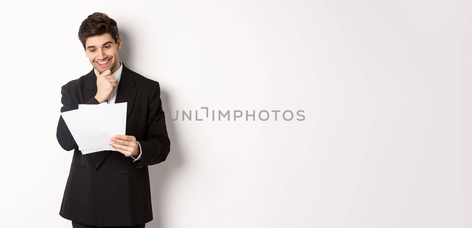 Image of handsome businessman in black suit, looking pleased at documents, reading report and smiling, standing against white background by Benzoix