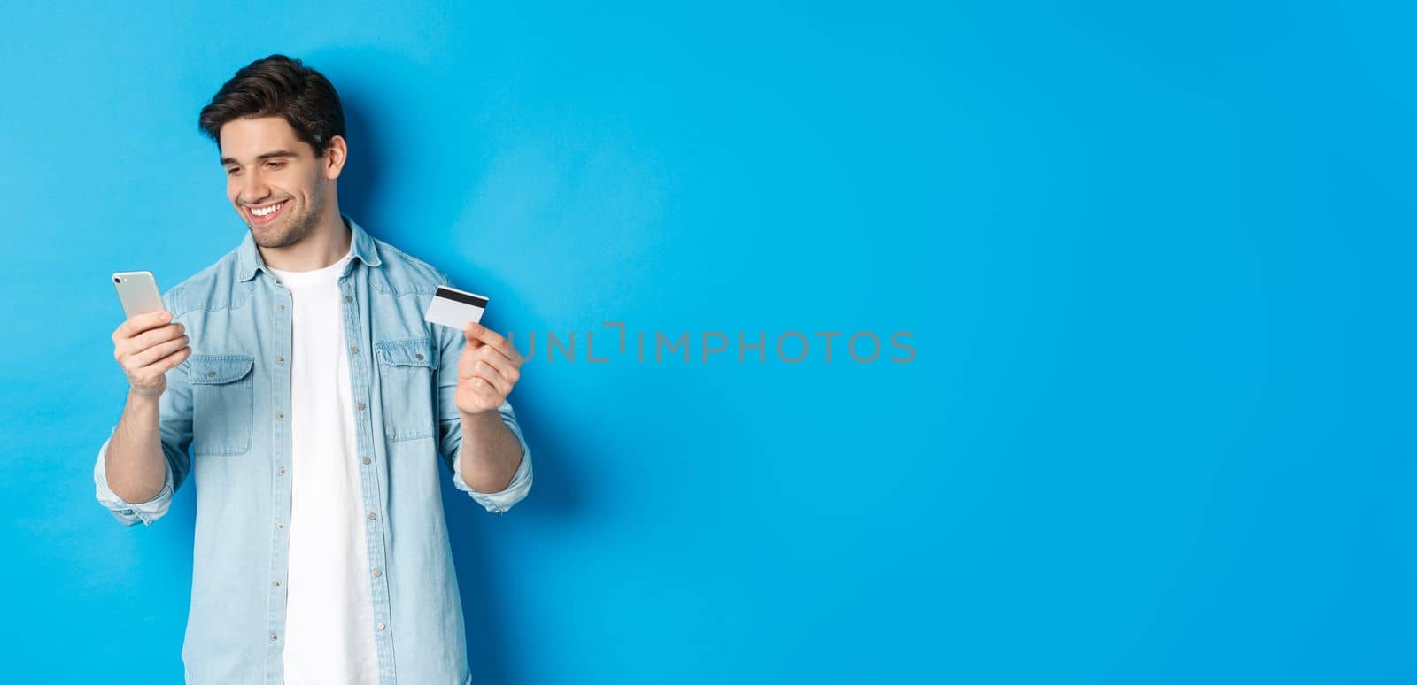 Young man shopping online with mobile application, holding smartphone and credit card, standing over blue background by Benzoix