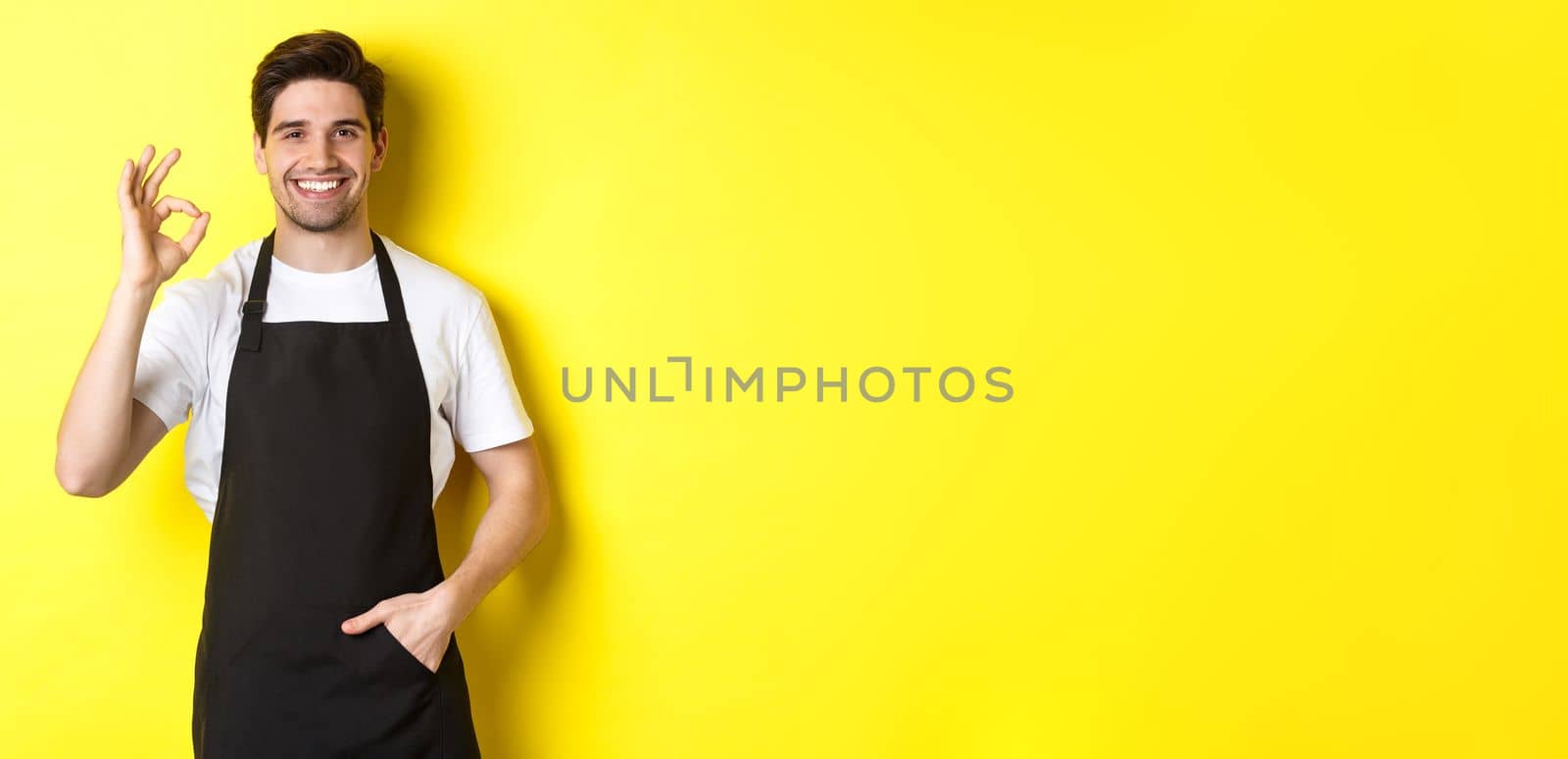 Young smiling barista in black apron showing okay sign, recommending coffee shop or restaurant, standing over yellow background by Benzoix