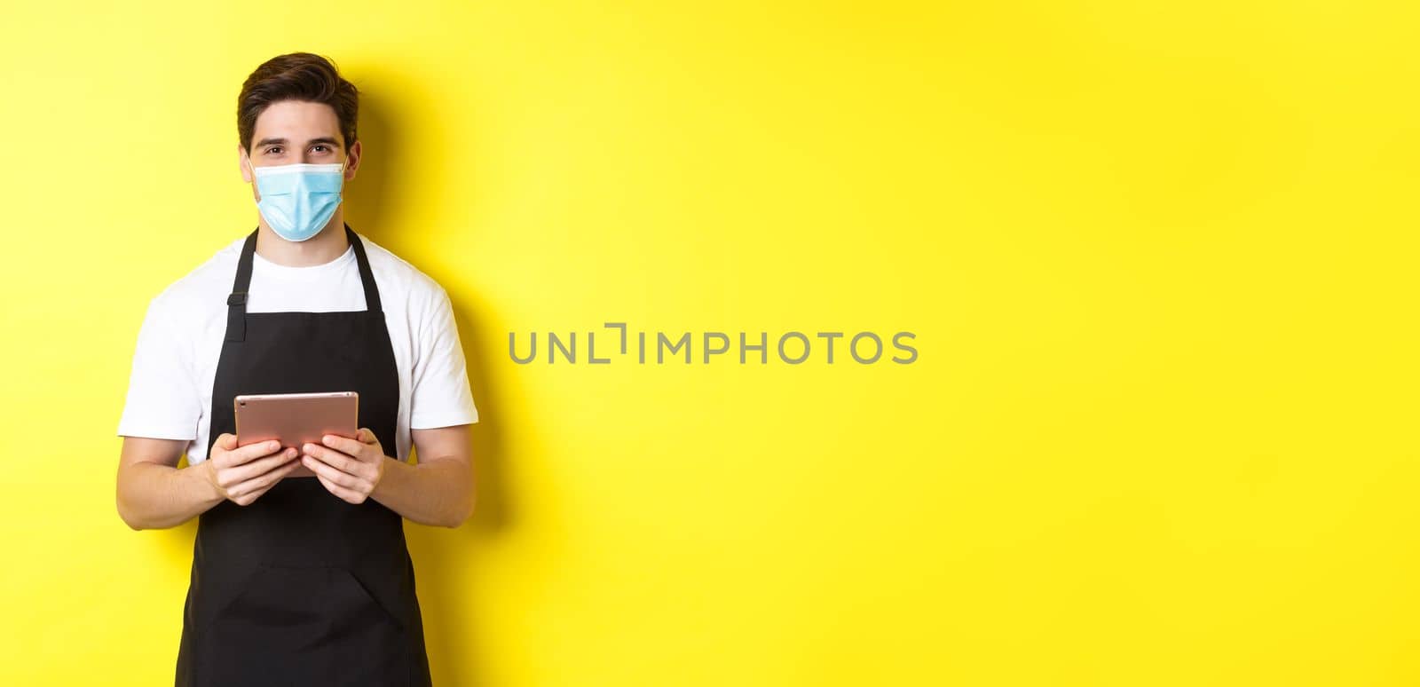 Concept of covid-19, small business and pandemic. Waiter in black apron and medical mask taking order, holding digital tablet, standing over yellow background by Benzoix