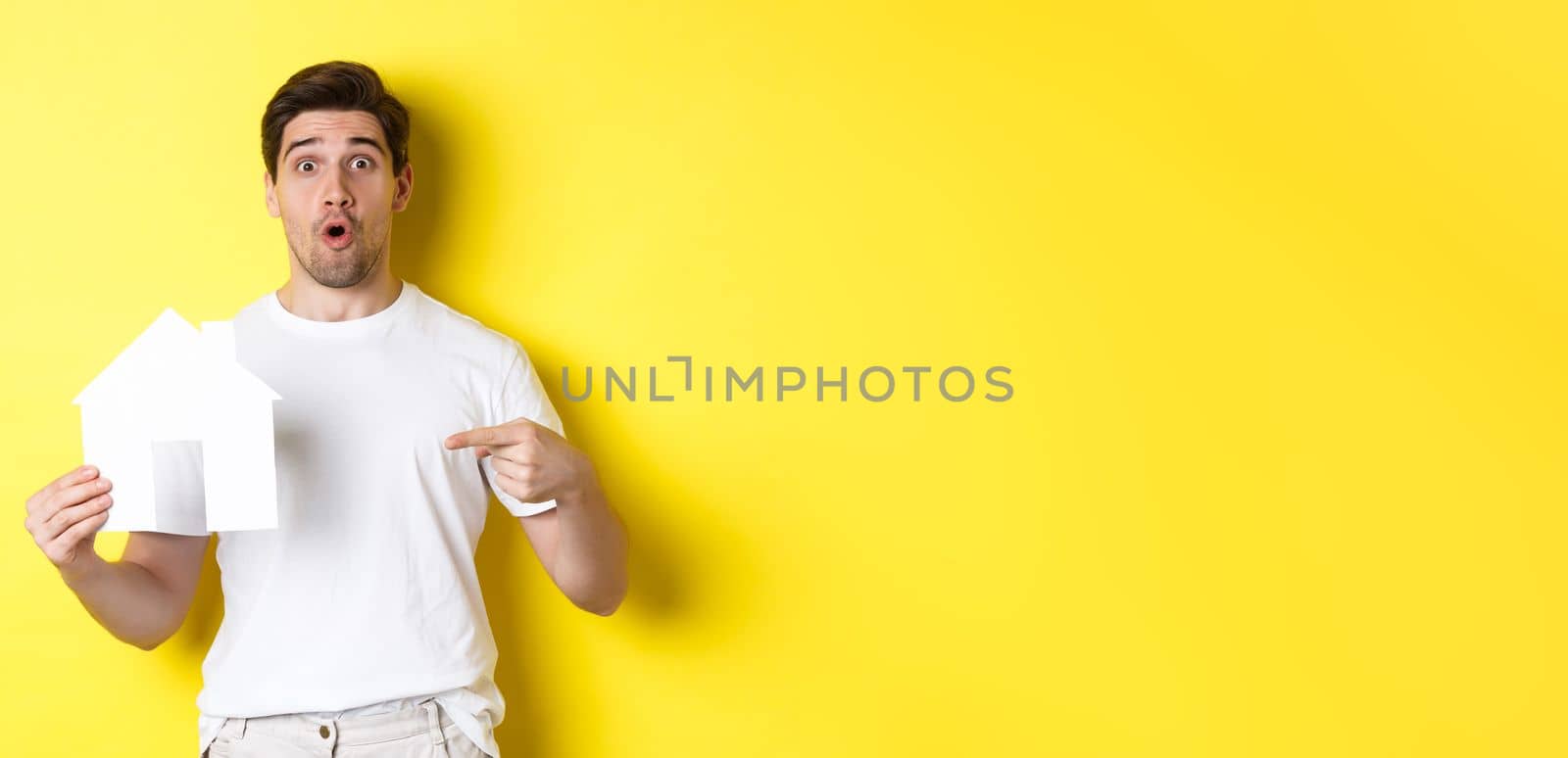 Real estate concept. Excited man pointing finger at paper house model, searching for flat, standing over yellow background by Benzoix