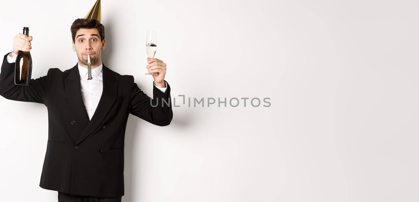 Concept of holidays and lifestyle. Handsome guy celebrating birthday, blowing party whistle and holding champagne, saying a toast, standing in suit over white background by Benzoix