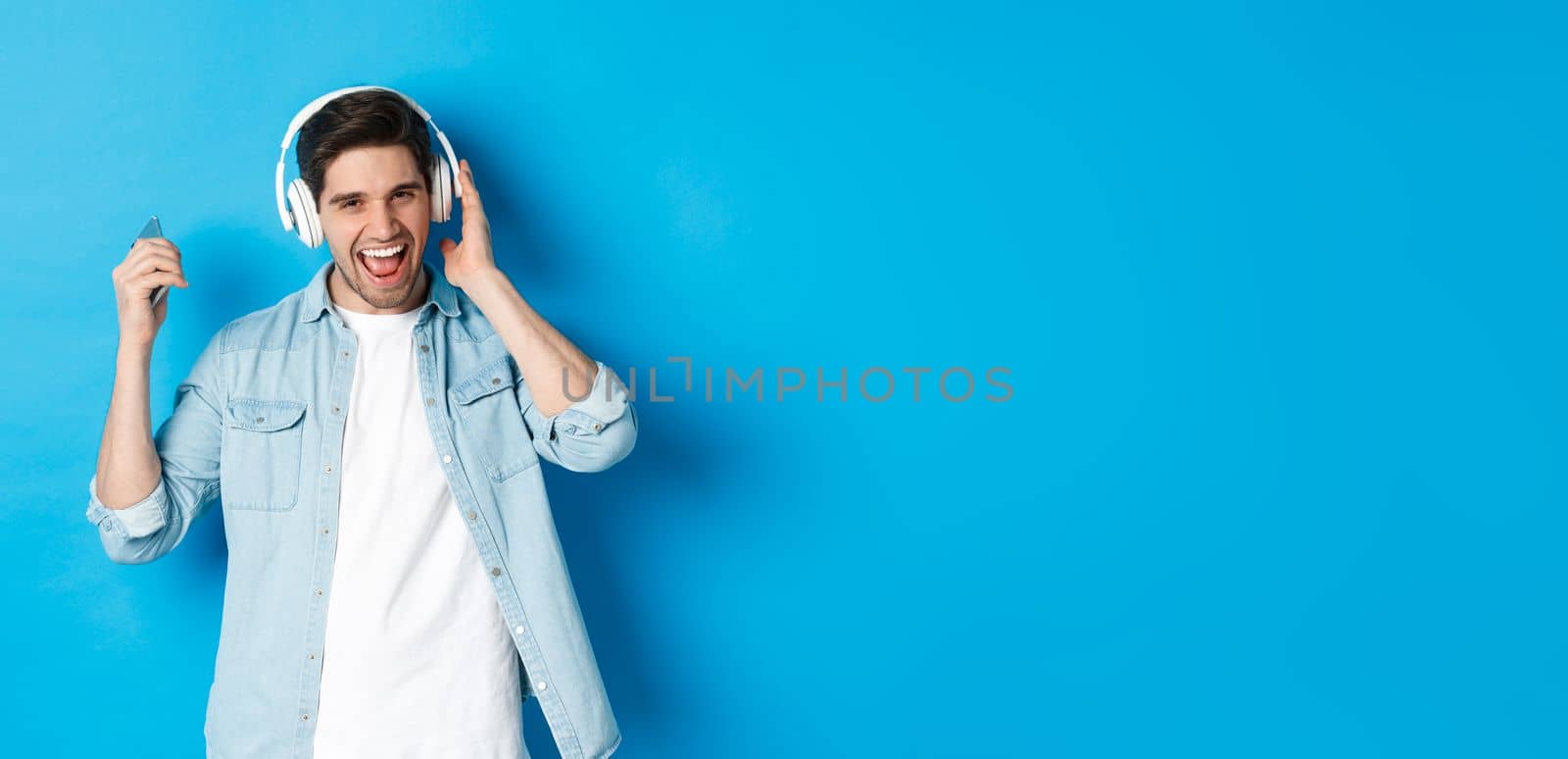 Happy smiling man enjoying listening to music in headphones, holding smartphone in raised hand, standing over blue background.