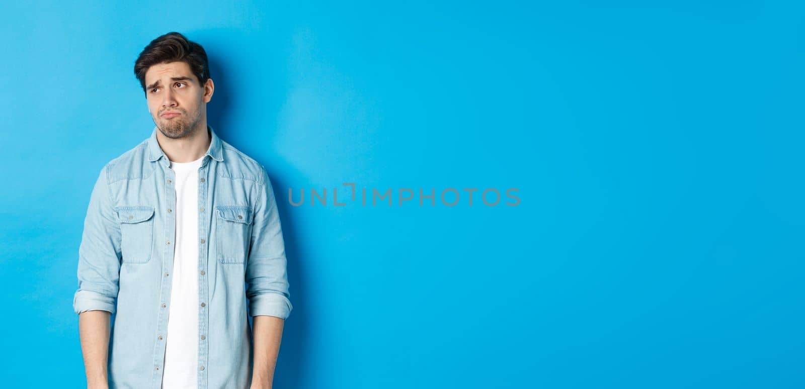 Image of reluctant and sad guy in casual outfit looking left, frowning and feeling upset, standing against blue background.