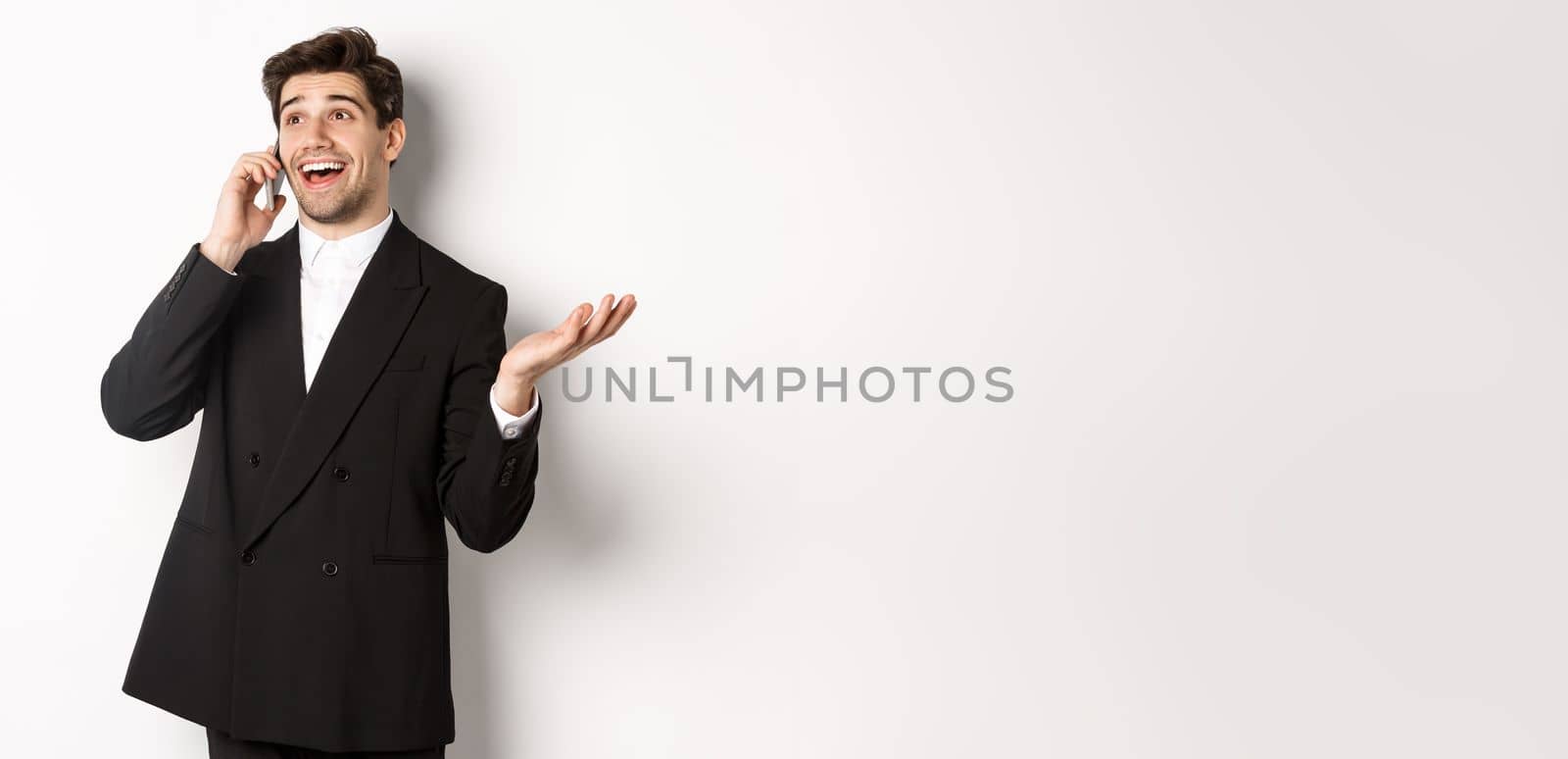 Portrait of happy good-looking businessman receiving great offer, talking on phone and looking pleased, standing in black suit against white background.