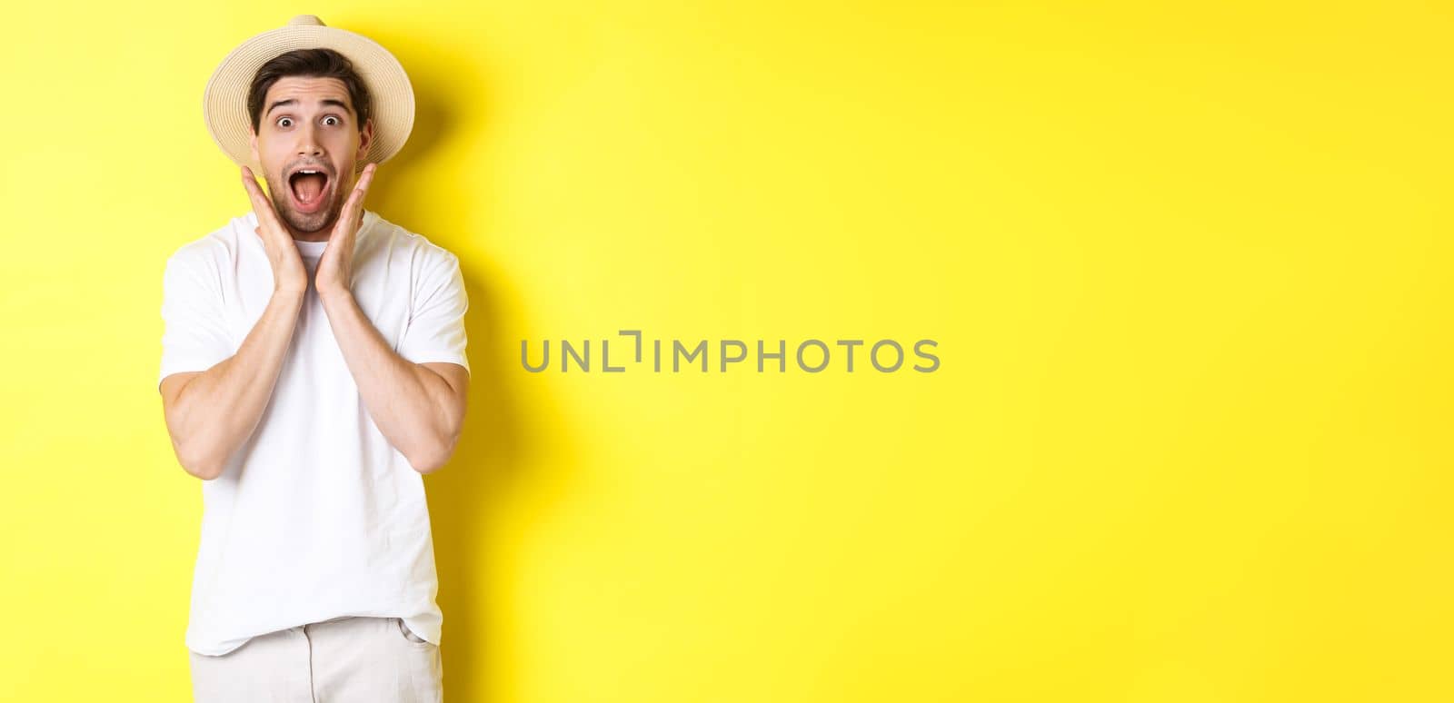 Concept of tourism and summer. Surprised male model in straw hat, looking amazed at special offer, standing against yellow background.