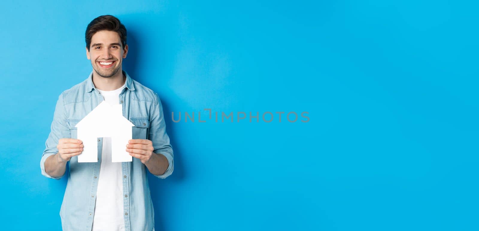 Insurance, mortgage and real estate concept. Smiling young man holding house model, searching apartment for rent, standing against blue background.