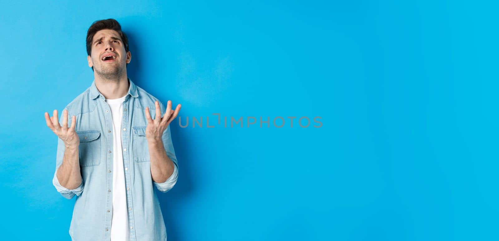 Frustrated guy complaining to god, looking up and pleading, standing against blue background upset.