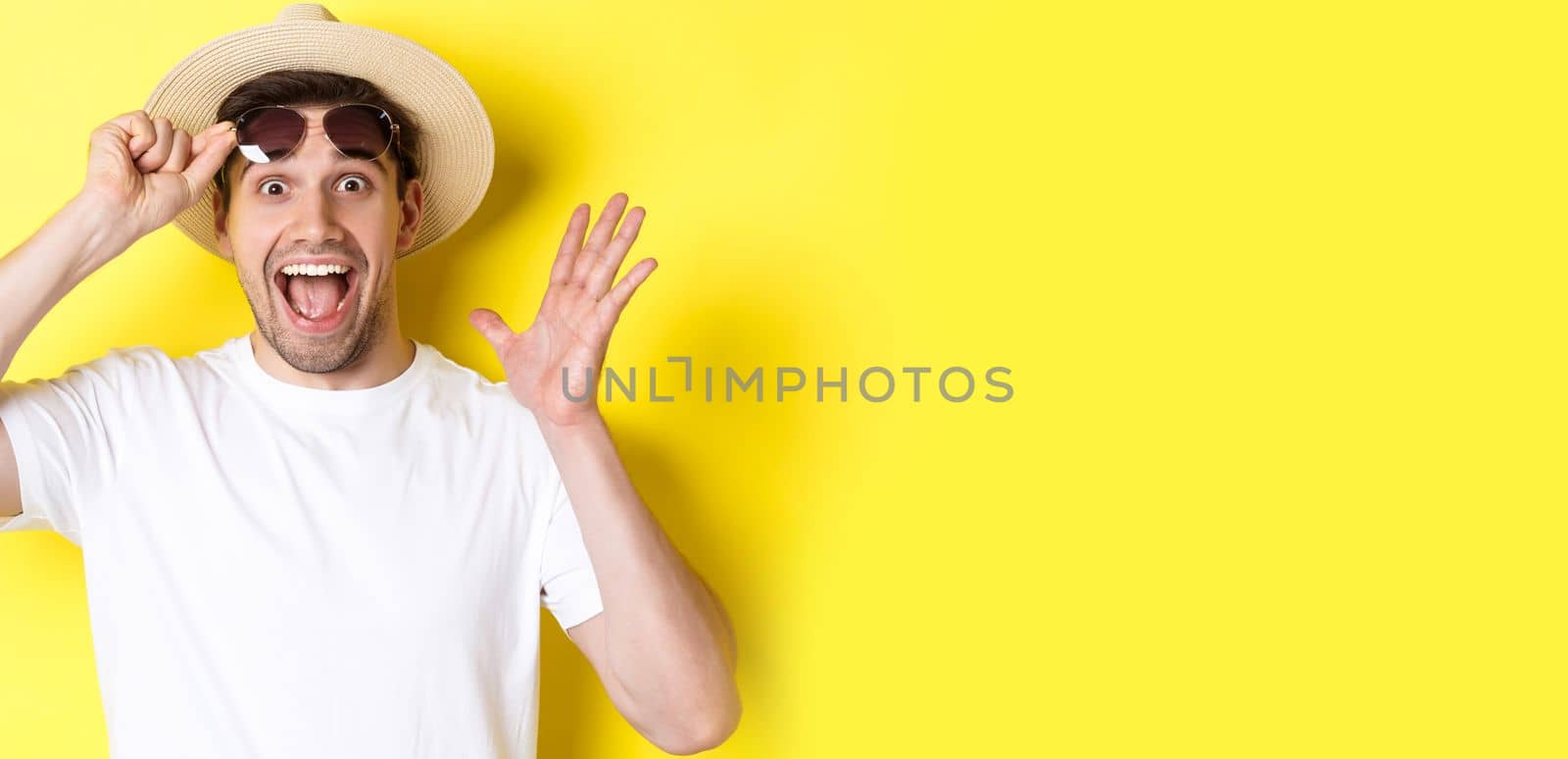 Concept of tourism and holidays. Excited man tourist take-off sunglasses and looking amazed at promo offer, standing over yellow background by Benzoix