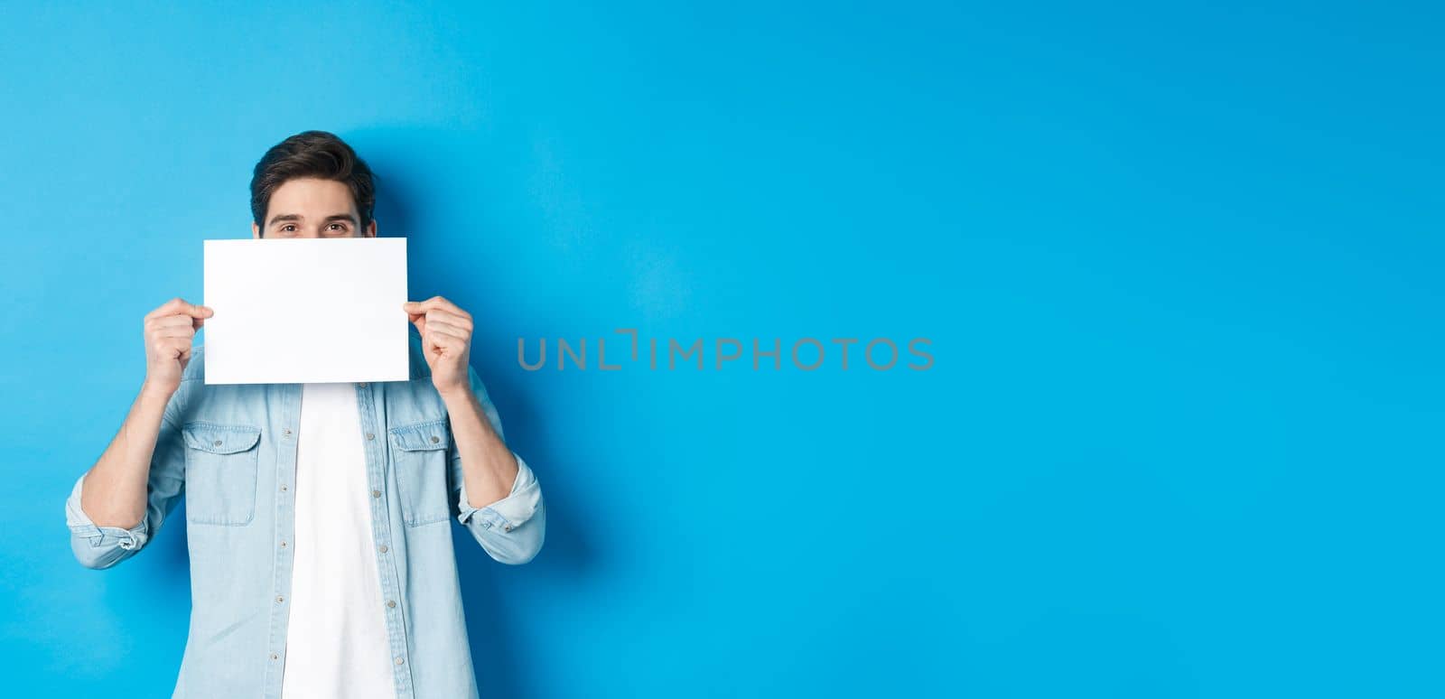 Sneeky handsome guy hiding face behind blank piece of paper for your logo, making announcement or showing promo offer, standing over blue background by Benzoix