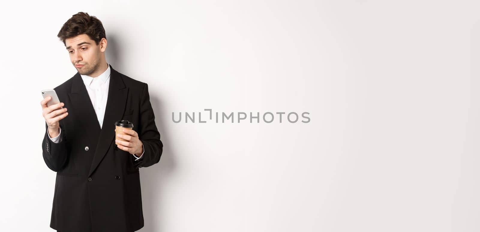 Portrait of thoughtful handsome businessman, drinking coffee and browsing in internet, looking at smartphone screen, standing against white background.