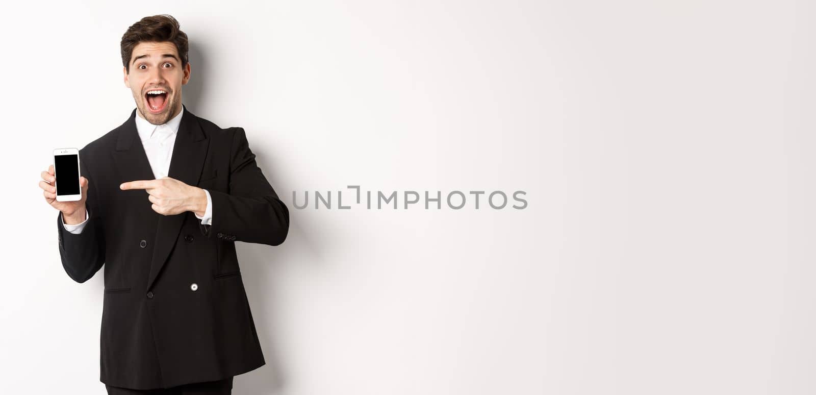 Portrait of handsome businessman in suit, pointing finger at mobile phone screen, showing advertisement, standing over white background.