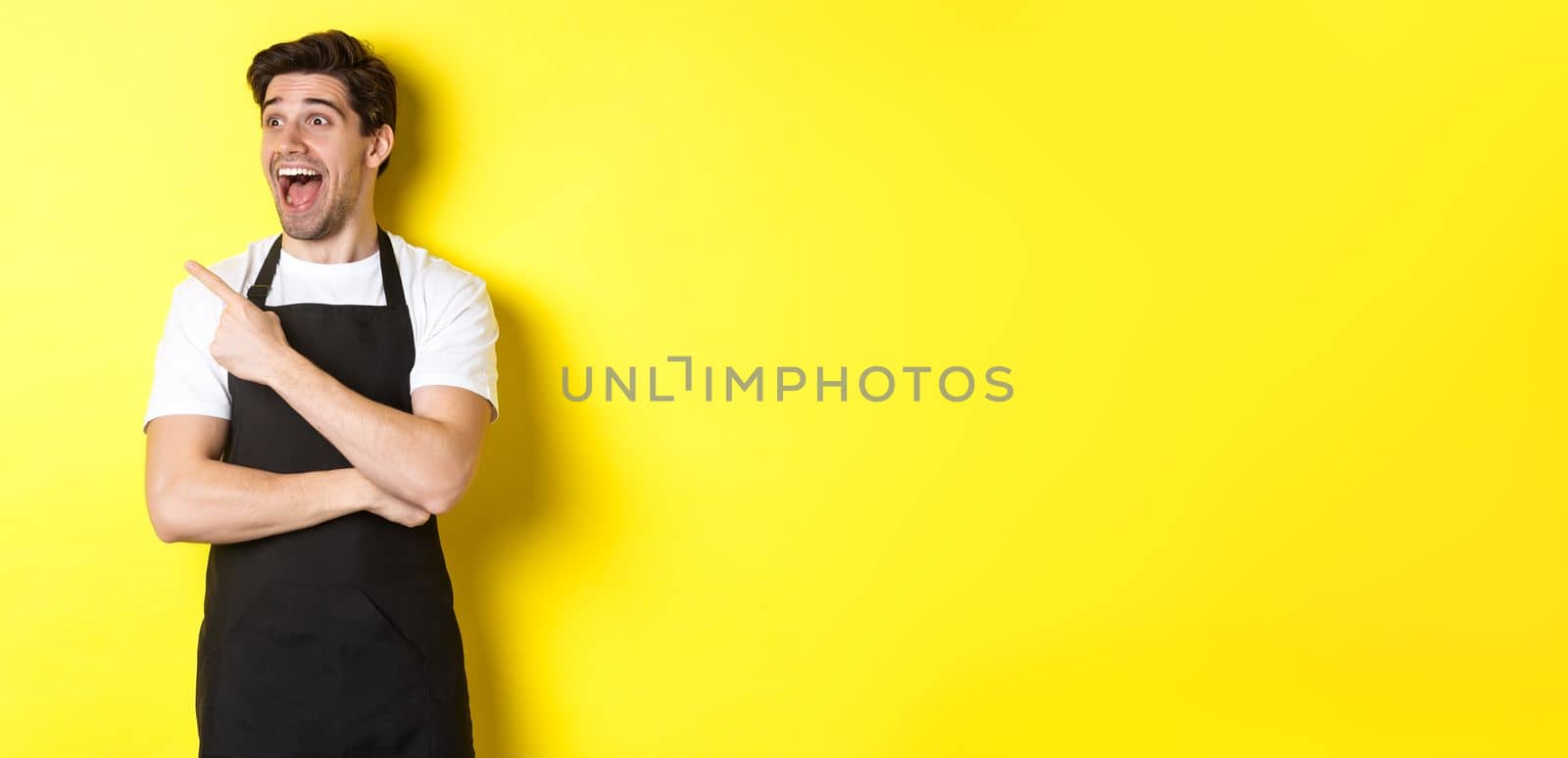 Waiter in black apron checking out promo offer, pointing finger and looking left at your logo, standing over yellow background.