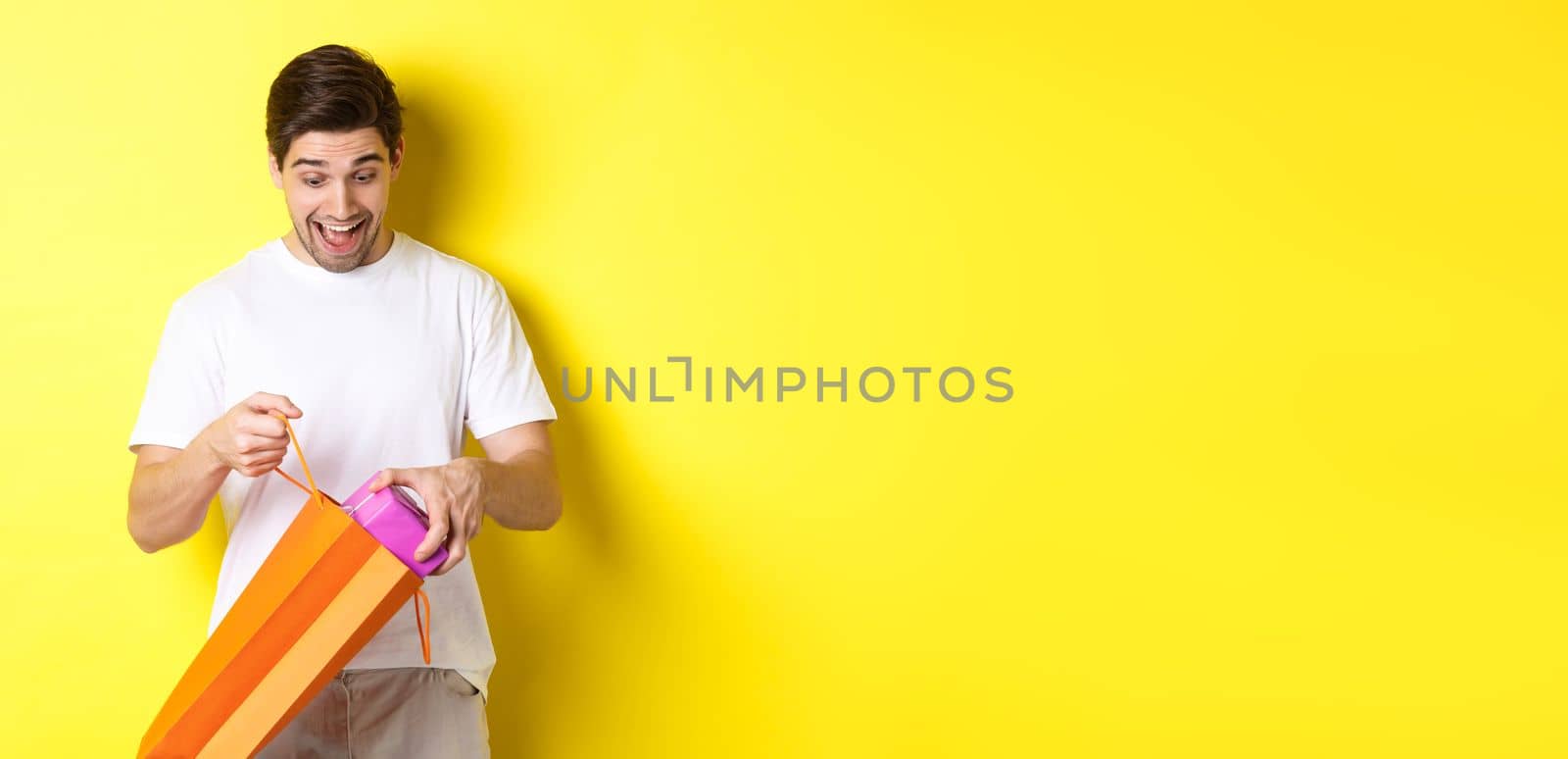 Concept of holidays and celebration. Young man looking surprised as take out gift from shopping bag, standing over yellow background by Benzoix