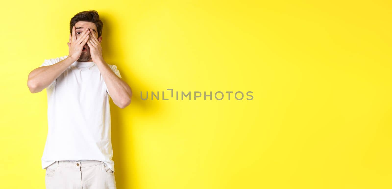 Embarrassed guy shut eyes but peeking through fingers at something awkward, standing over yellow background by Benzoix